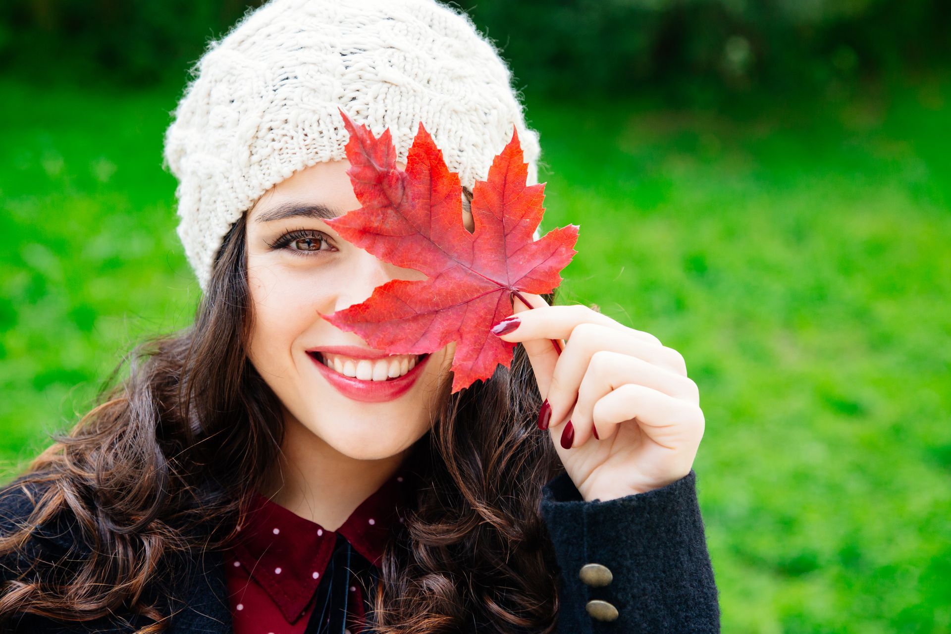 Woman smiling, holding red maple leaf over one eye, wearing white knit hat, outdoors.