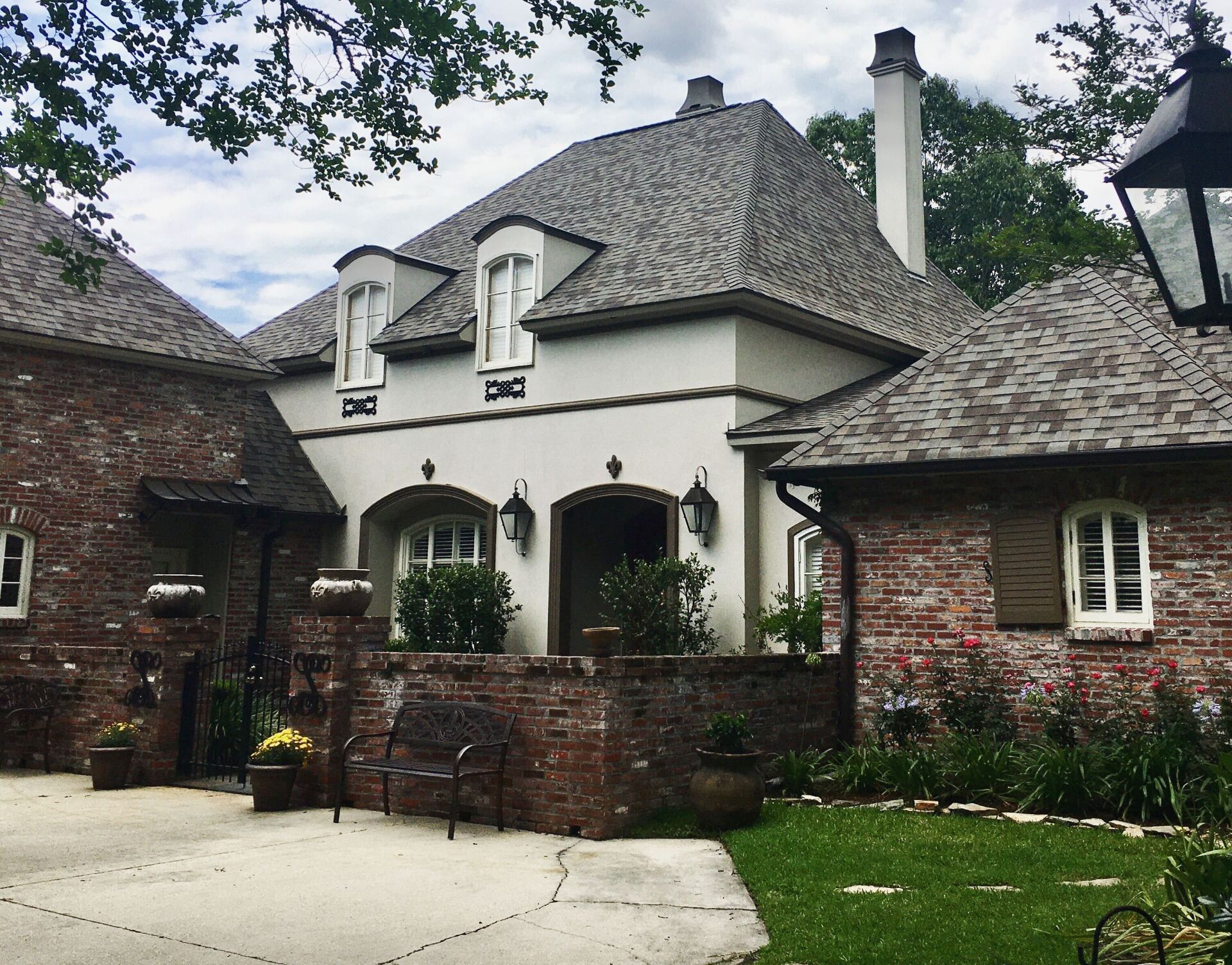 Brick and stucco home with a dark roof and multiple dormers. A brick wall encloses a patio area with flowers.