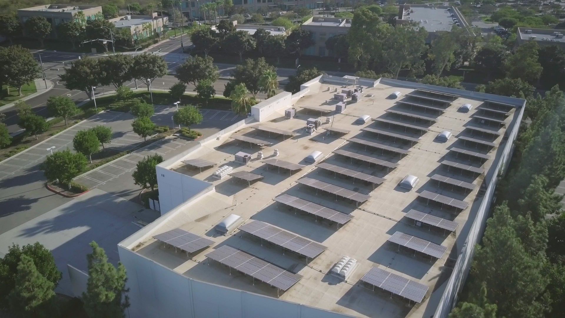 Aerial view of a flat building roof featuring rows of solar panel canopies, surrounded by trees and a parking lot.