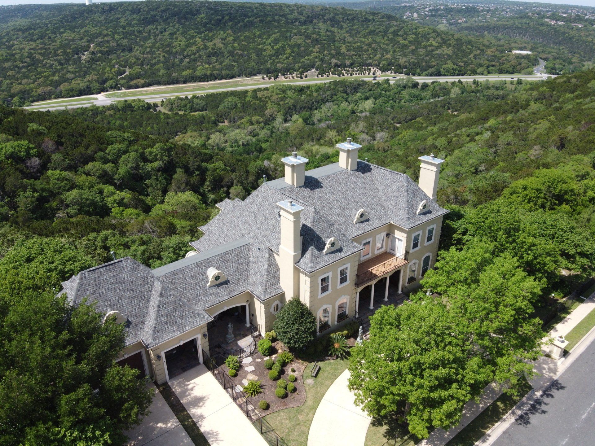 Large beige mansion with a gray roof nestled in a forested hillside.