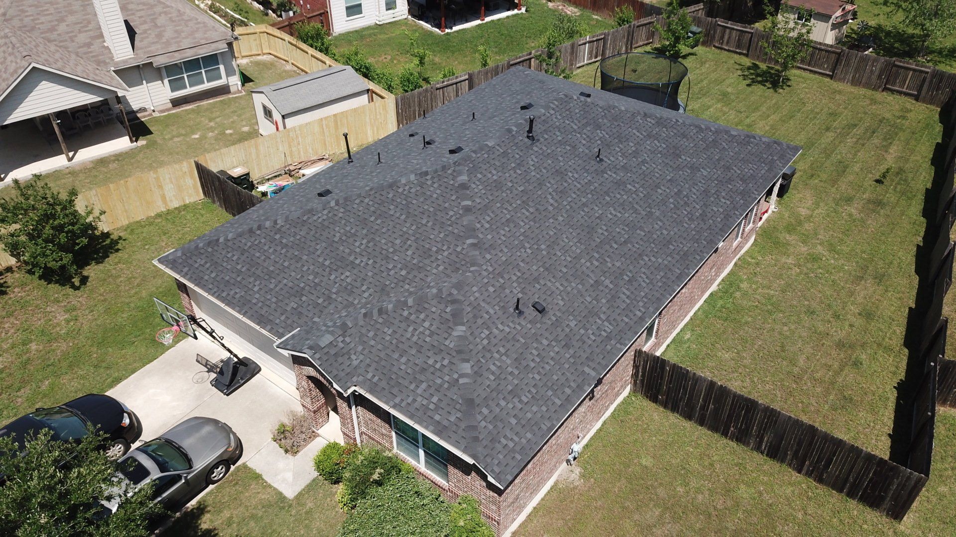 Aerial view of a house with a dark gray roof, surrounded by a fence and green lawn. Cars are parked in the driveway.