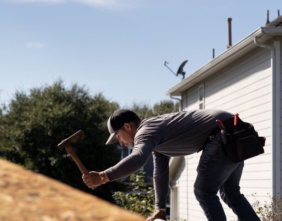 Roofer hammering on a roof near a white house with a satellite dish; sunny day.