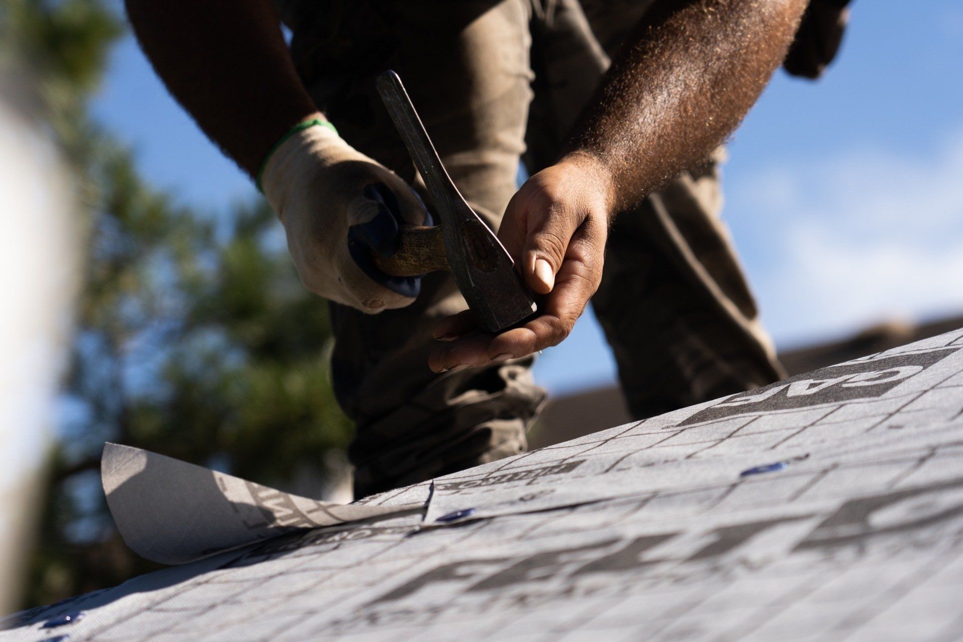 A roofer cutting roofing material on a rooftop; blue sky in background.