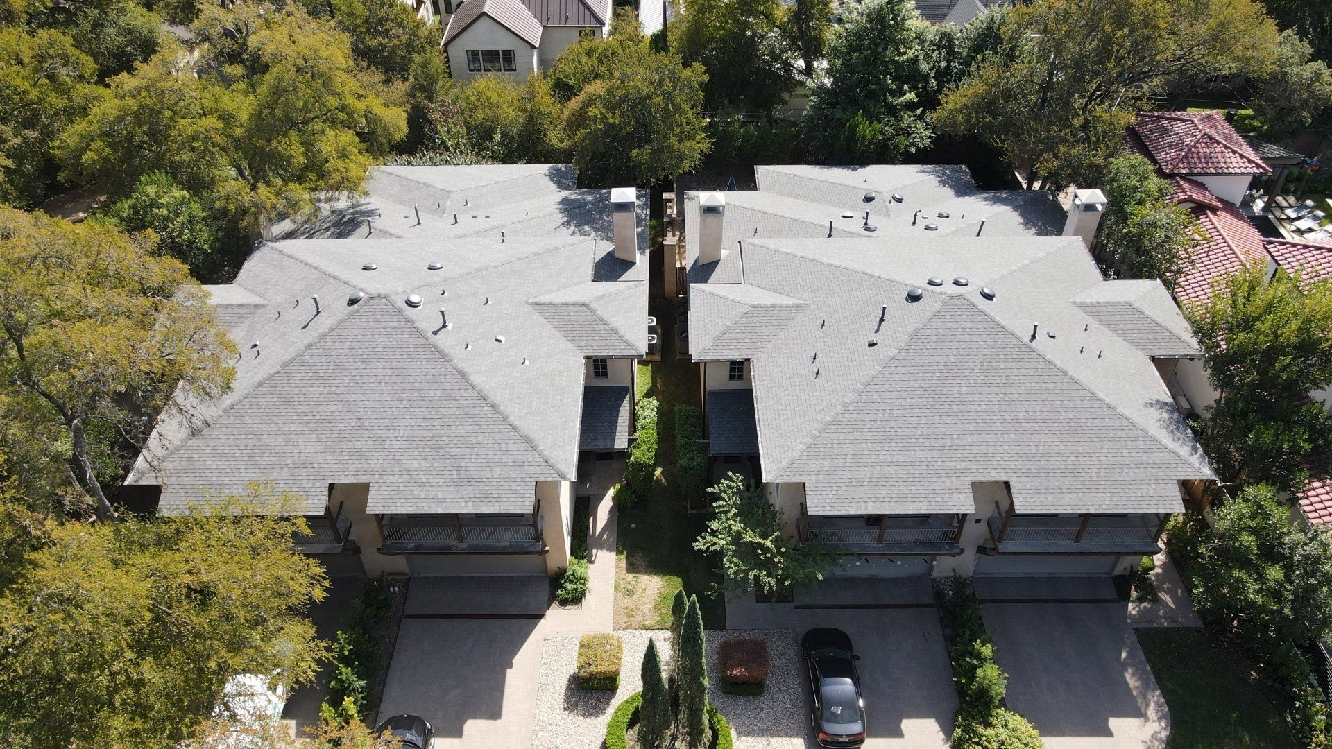 Aerial view of two connected gray-roofed townhouses with green trees and parked cars.