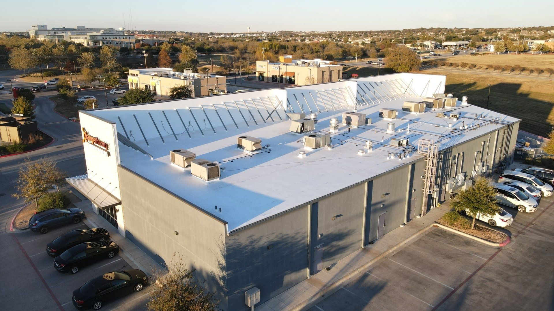 Exterior aerial view of a large industrial building with a white roof, and parked cars in the parking lot.