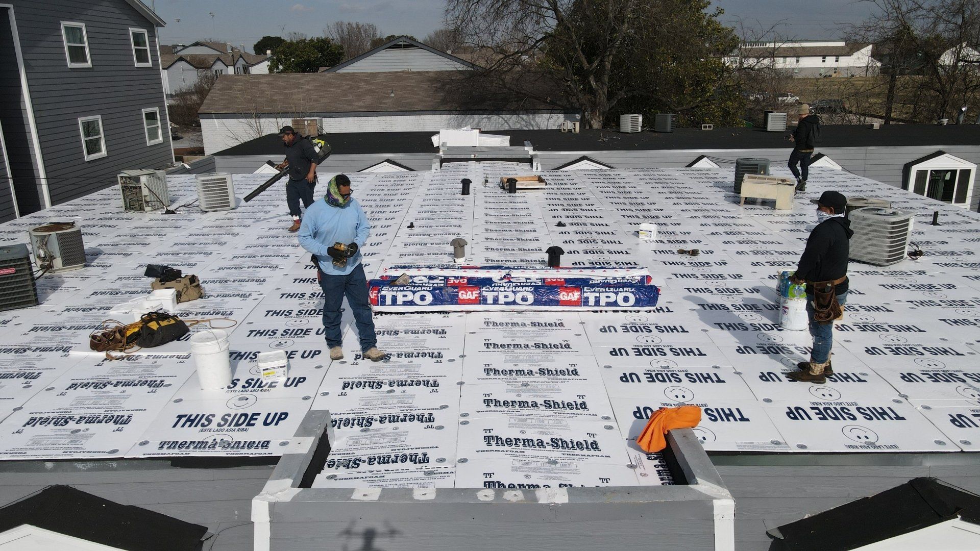 Roofers working on a flat roof, installing white roofing material, surrounded by equipment.