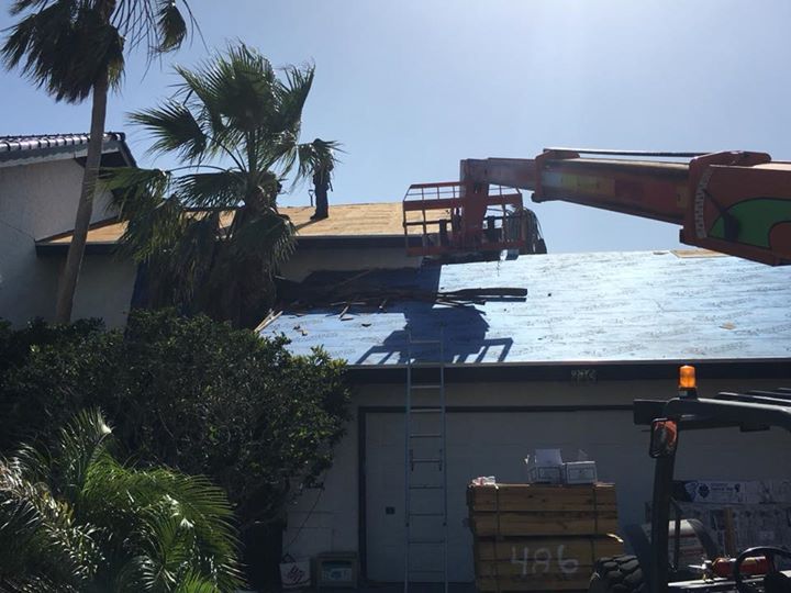 Roofing work in progress; an orange lift extends toward a house with a blue underlayment and partially removed roof.