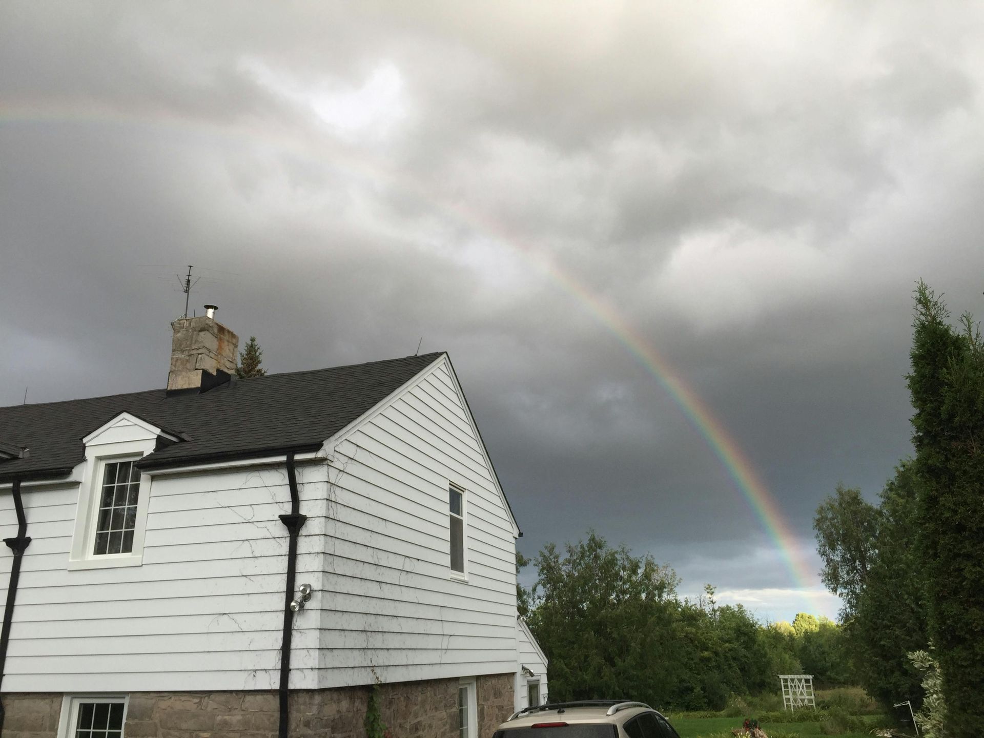 Rainbow arcs over a white house with dark roof against a cloudy sky.