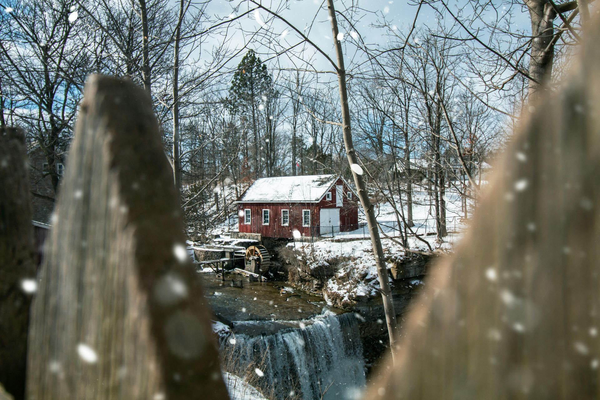 Snowy scene: Red building by a waterfall framed by a wooden fence. Bare trees, flakes falling.