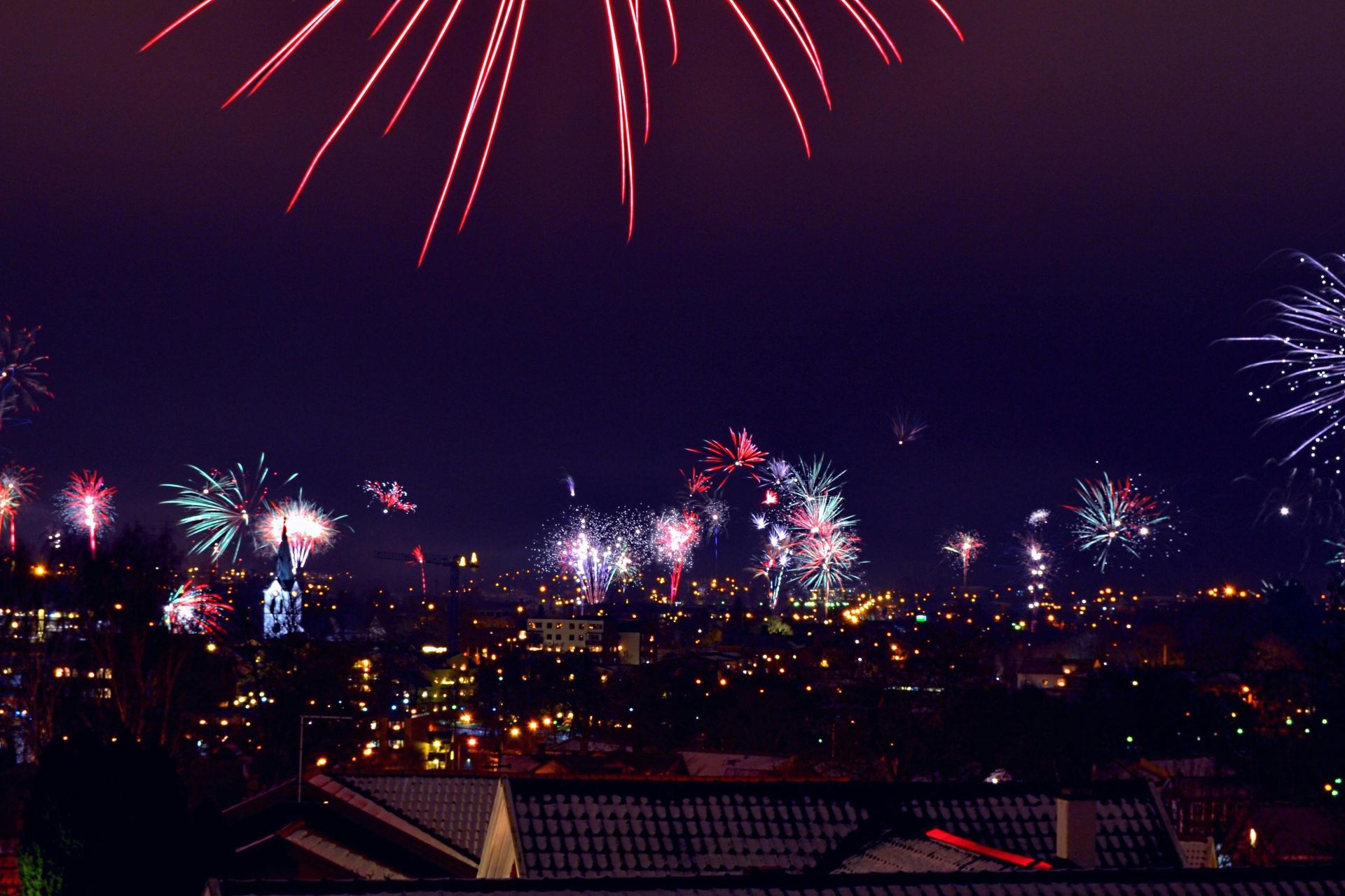 Fireworks exploding over a city at night, with red, blue, and white bursts against a dark purple sky.