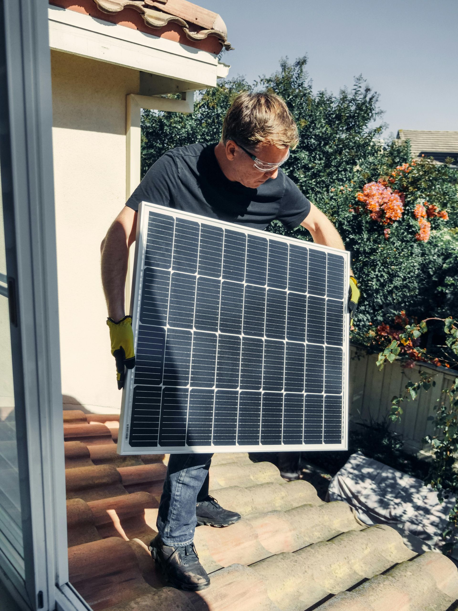 Man holding a solar panel on a tiled roof. He wears safety glasses and gloves, with a sunny backdrop.