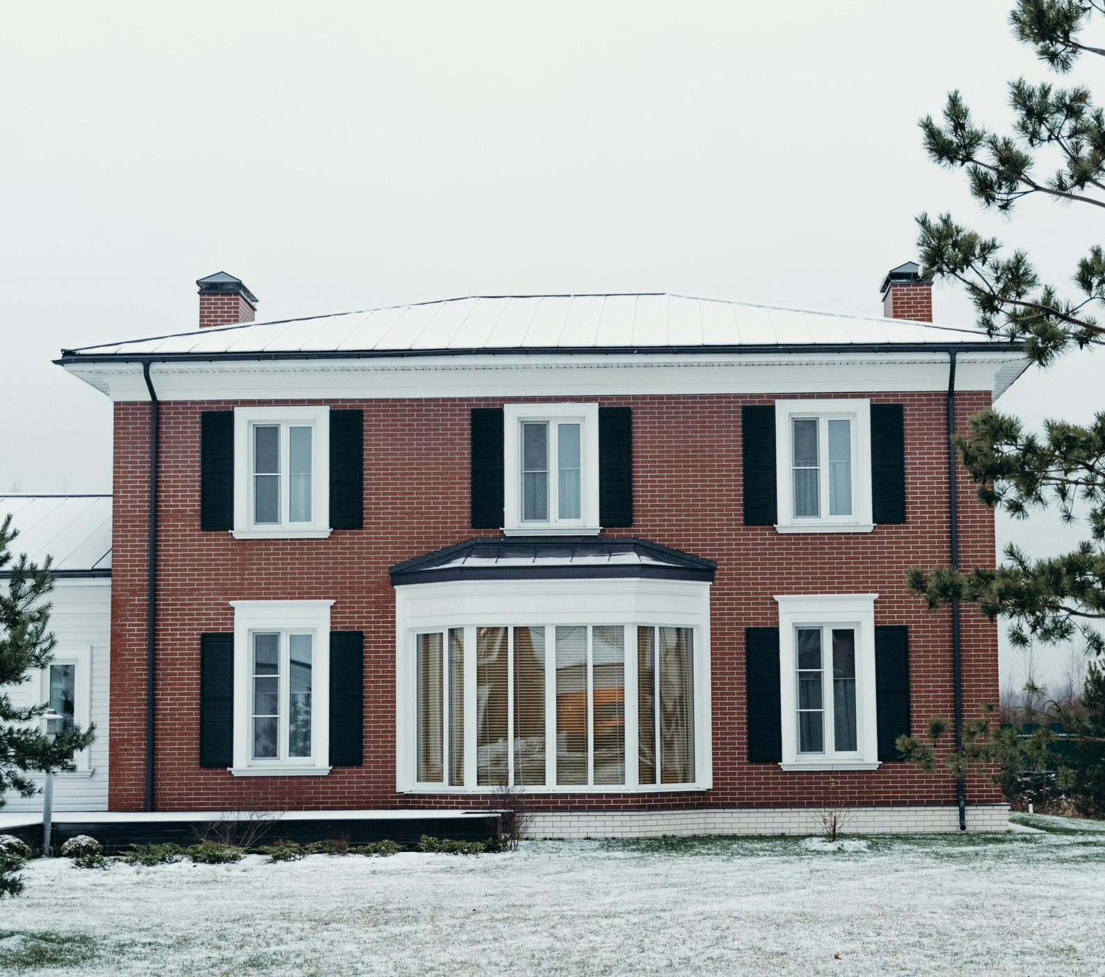 Brick house with snow-covered roof and lawn, black shutters, bay window, and a wintery backdrop.