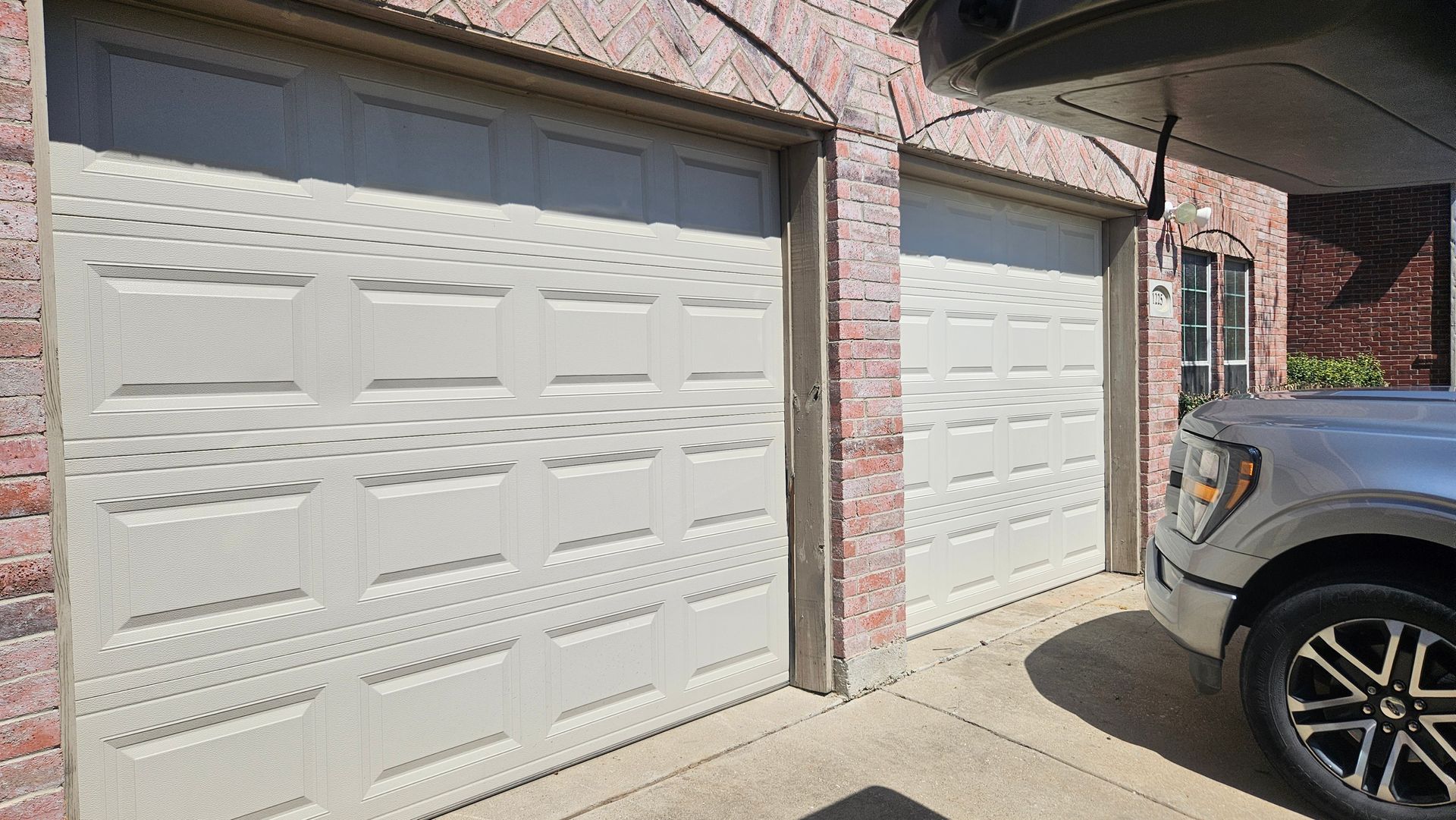 Two beige garage doors and a pickup truck parked outside, near a brick building.