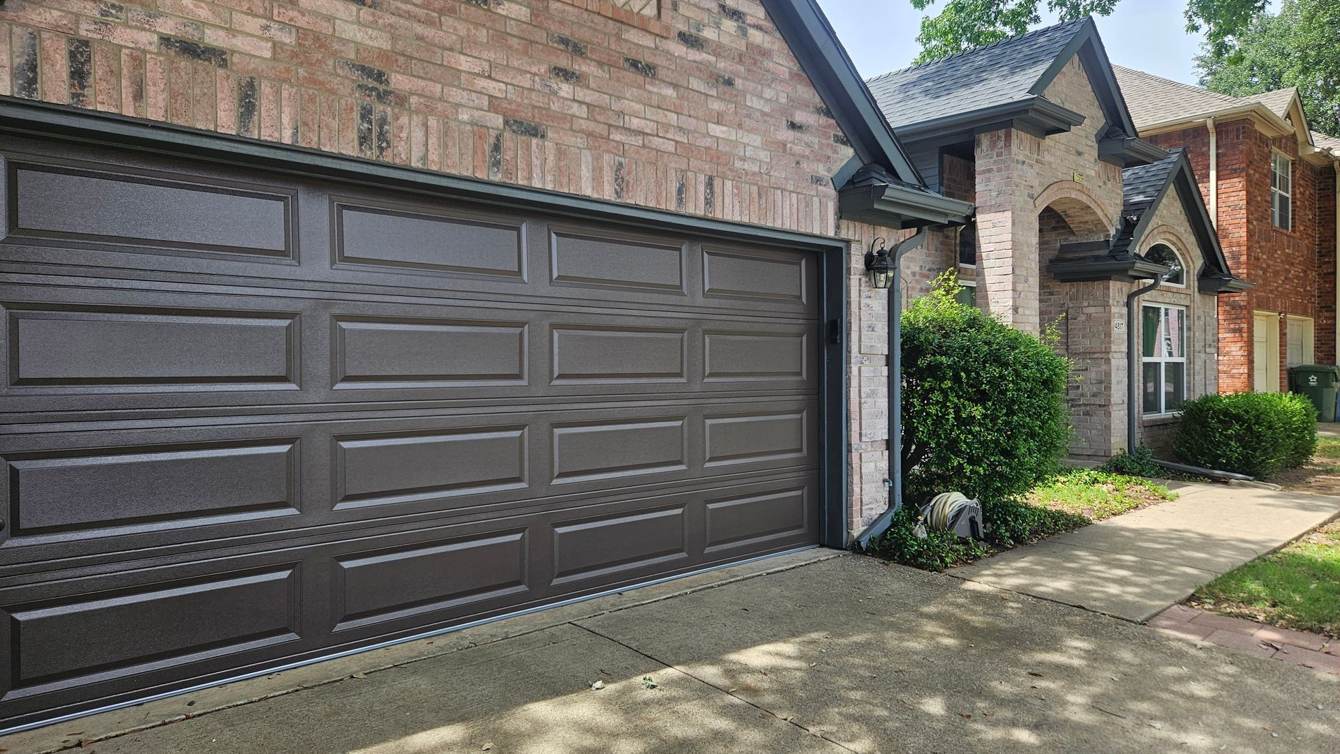 Brown garage door on a brick house with a walkway, green shrubs, and blue sky.