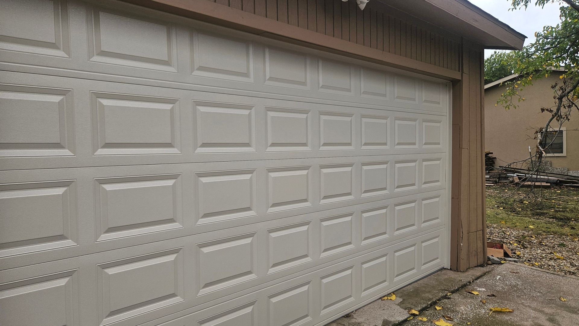 White garage door with grid pattern, brown trim, and partial view of a building in the background.