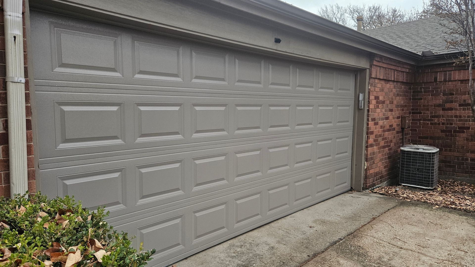 Gray garage door on a brick building with a gutter and trash can nearby.