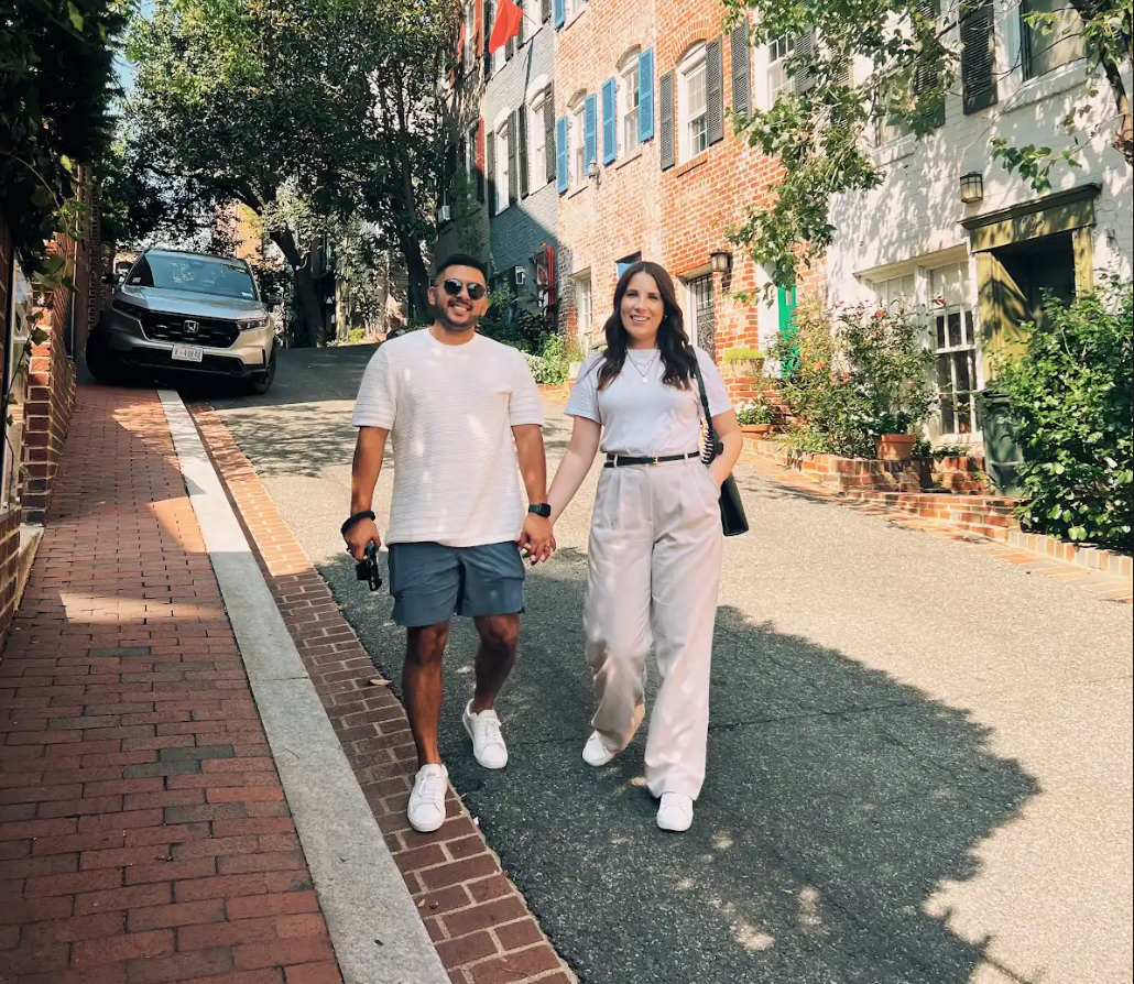 Two people walking hand in hand on a sunny cobblestone street, with brick buildings and a parked car nearby.