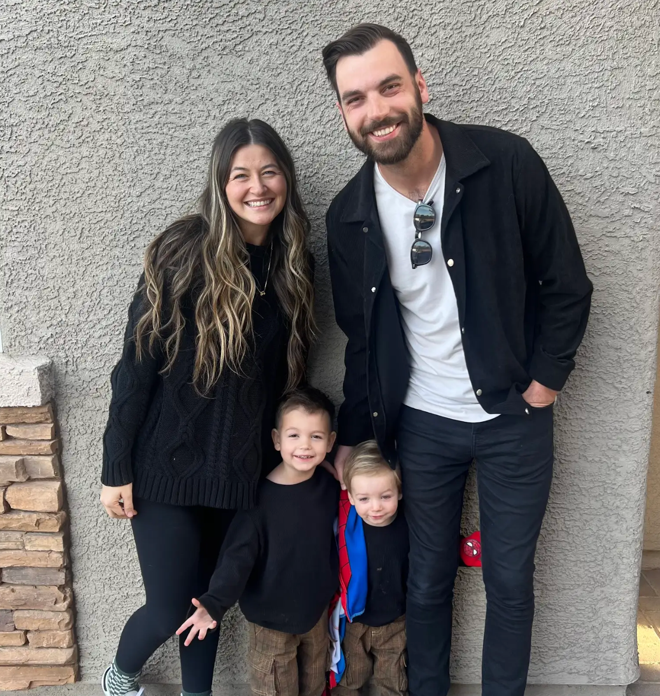 Smiling family of four standing against a textured wall, two adults behind two children.