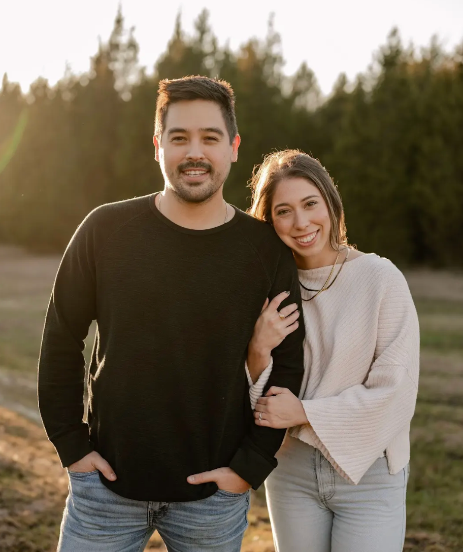 Two people standing outdoors, smiling, with trees in the background.