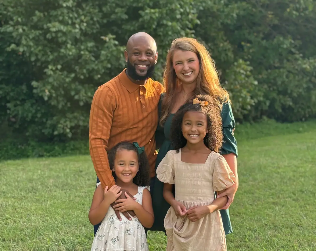 Family of four smiling outdoors in a grassy field, with trees in the background.
