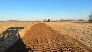 A dirt road going through a field with a shadow of a tractor on the ground.