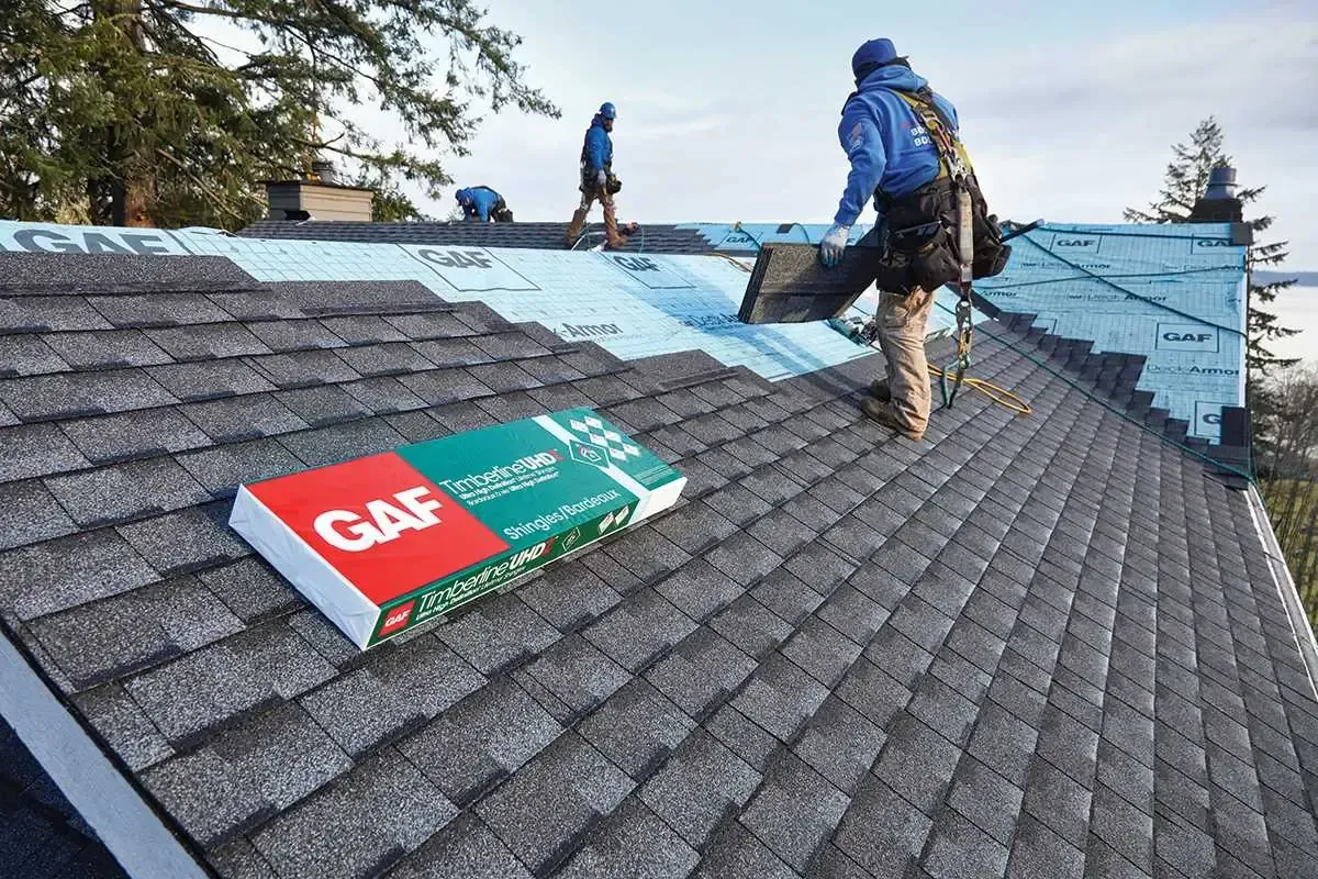 A man is standing on top of a roof with a box of gaf shingles.