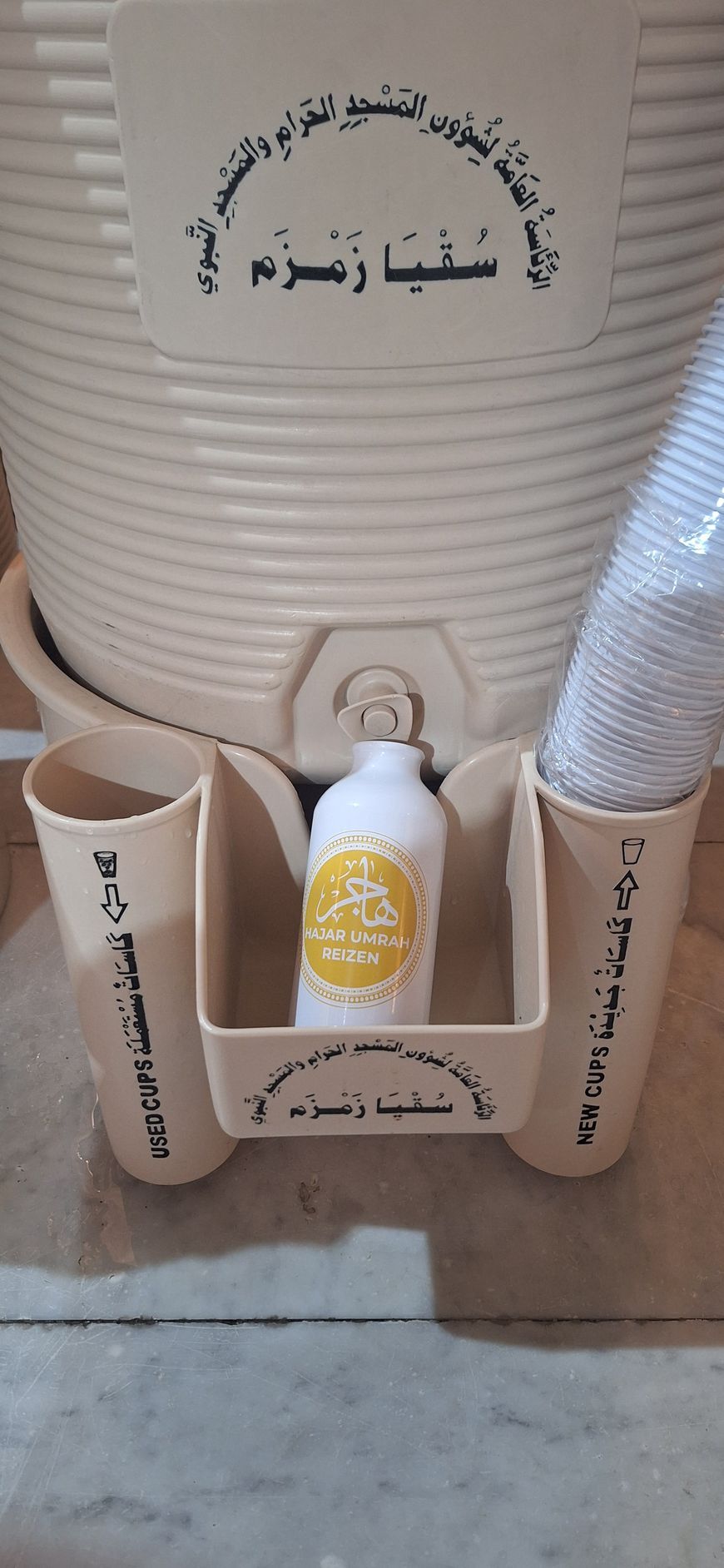 Een fles met vloeistof staat in een houder naast twee kopjes.