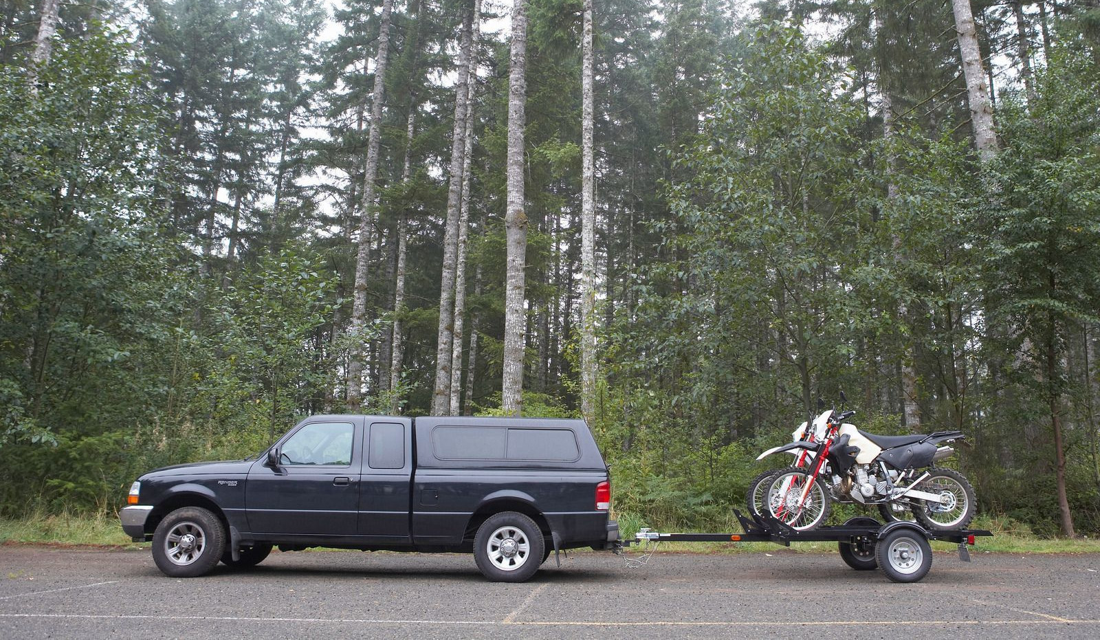 Dark truck towing a trailer with two motorcycles parked on the side of a road near a forest.