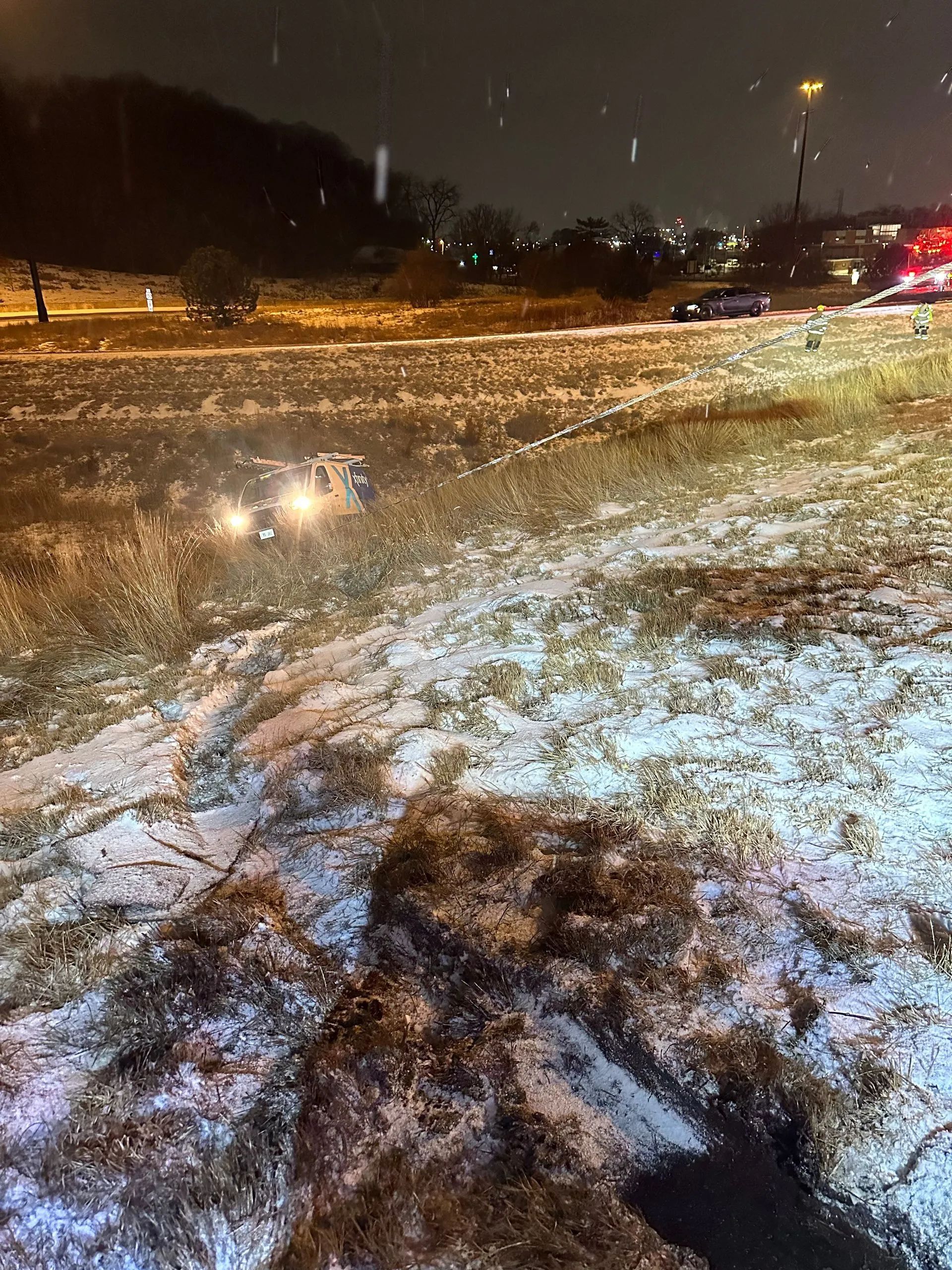 Snowy roadside scene at night with vehicle headlights, grassy area, and streetlights.