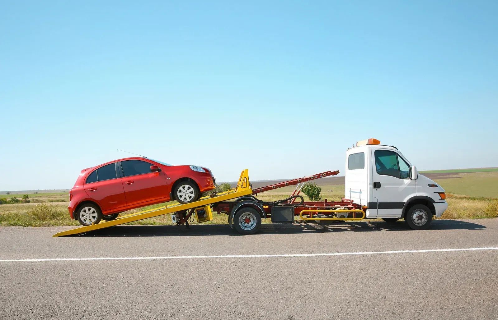 A red car is being towed by a tow truck on the side of the road.