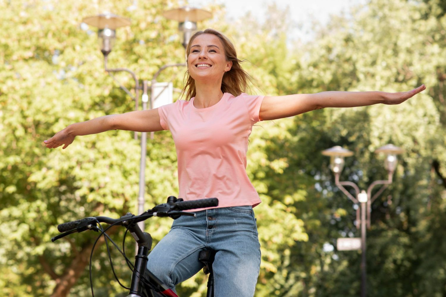 Woman riding a bicycle with arms outstretched, smiling in a park setting.