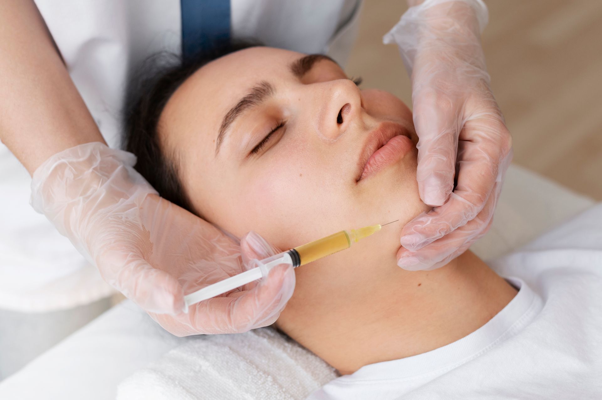 Woman receiving facial treatment with injection to the jawline, by a gloved person in a clinical setting.