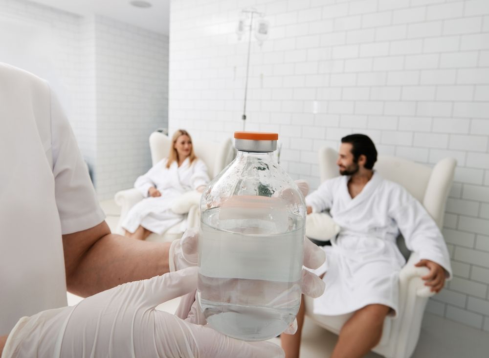 A healthcare worker holds IV fluids, with two people in robes in the background, sitting in white chairs.