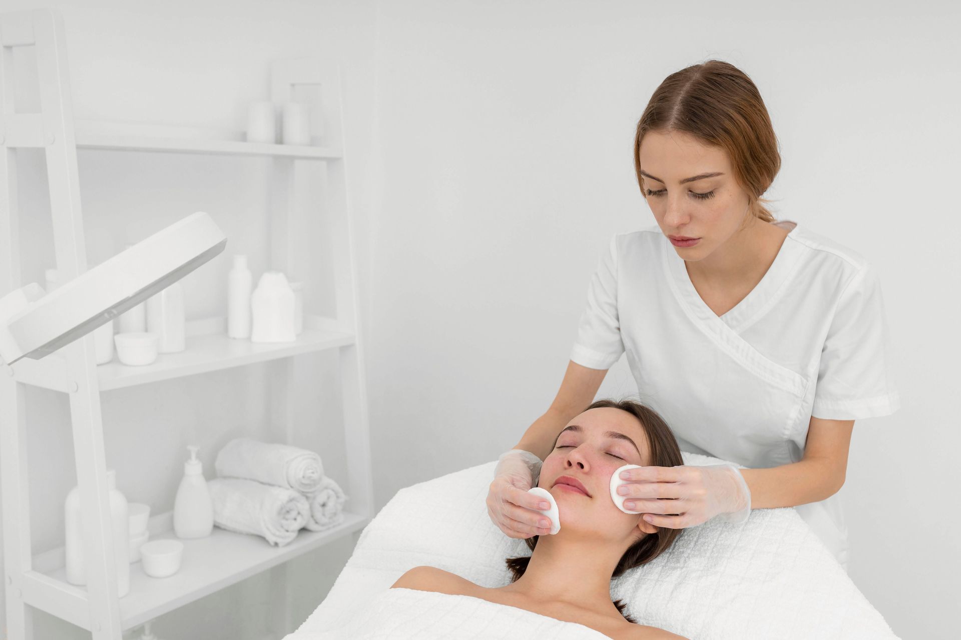 Woman receiving facial treatment at a spa. Therapist uses cotton pads. Bright, white room.