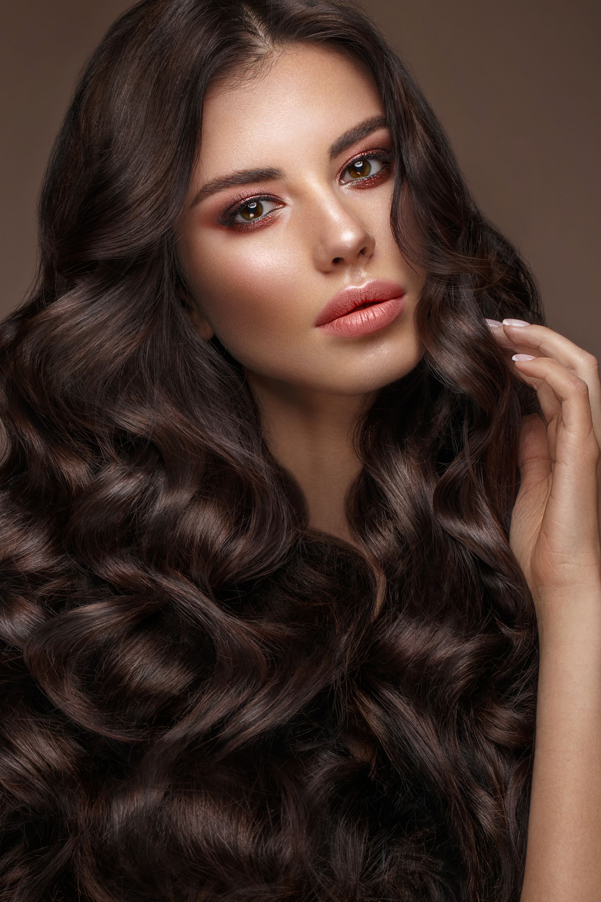 Woman with long, wavy dark brown hair, rosy cheeks, and full lips, hand touching hair.