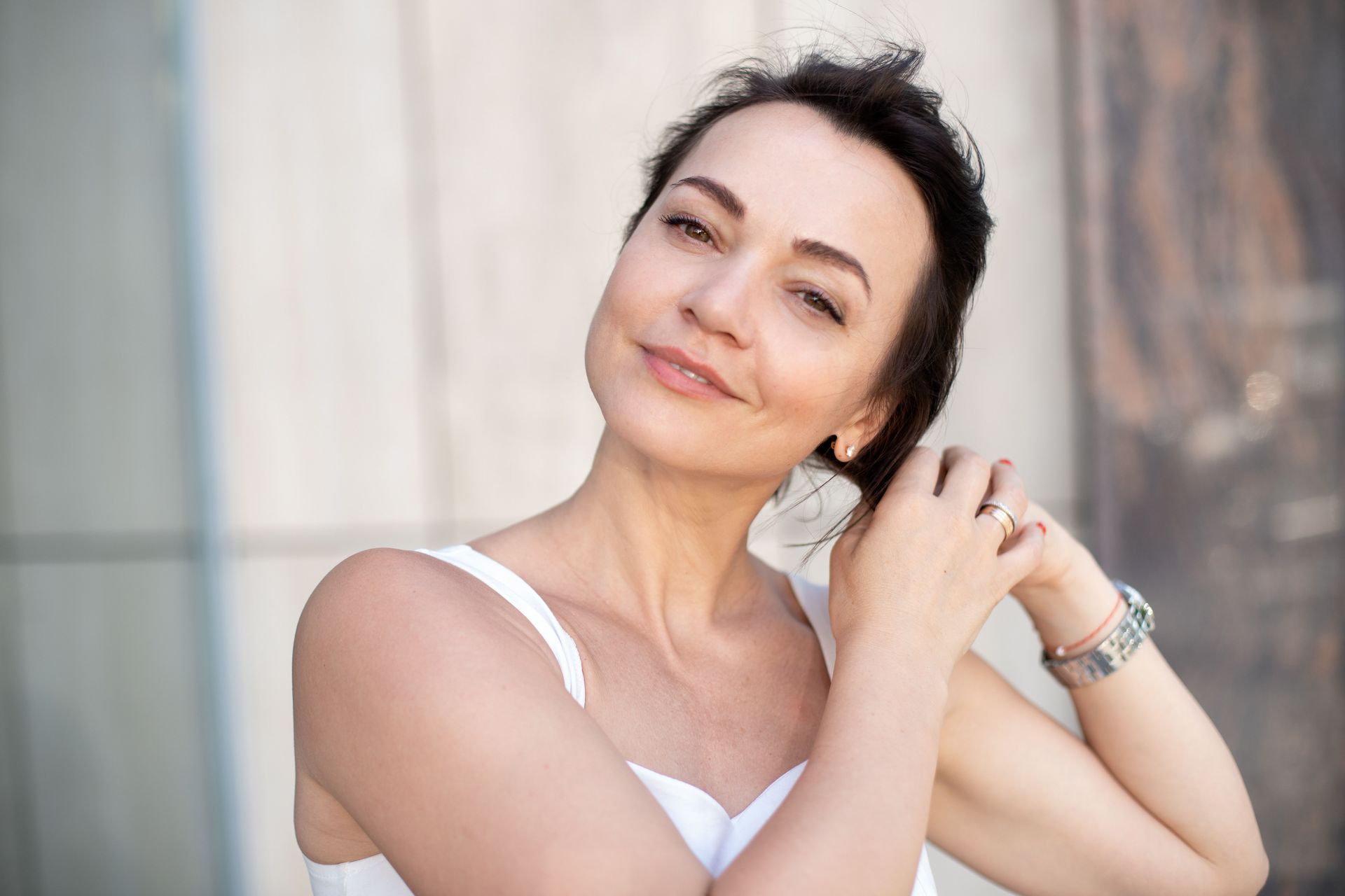 A woman in a white tank top is holding her hair and smiling.