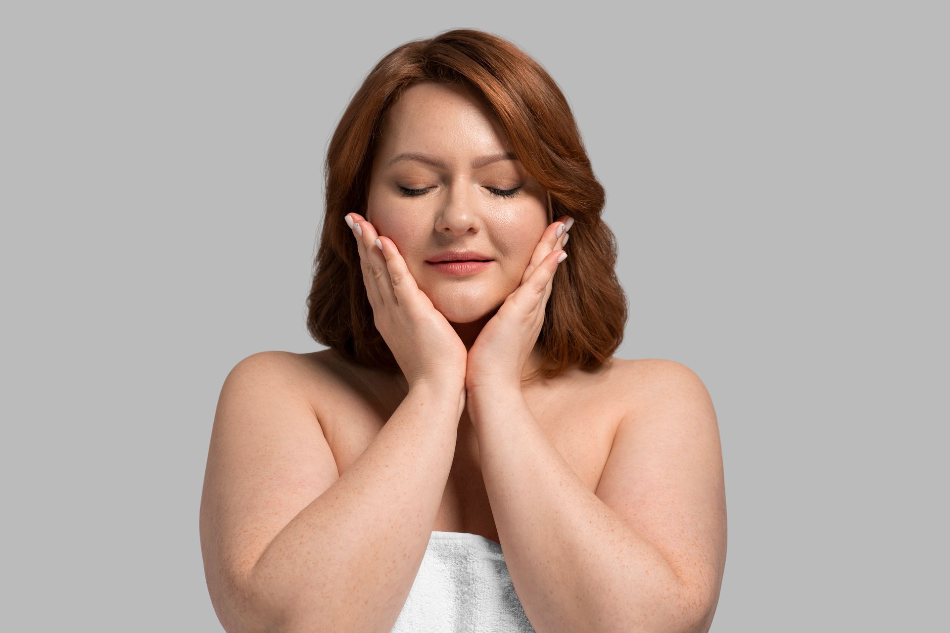 A woman is wrapped in a towel and touching her face with her hands.