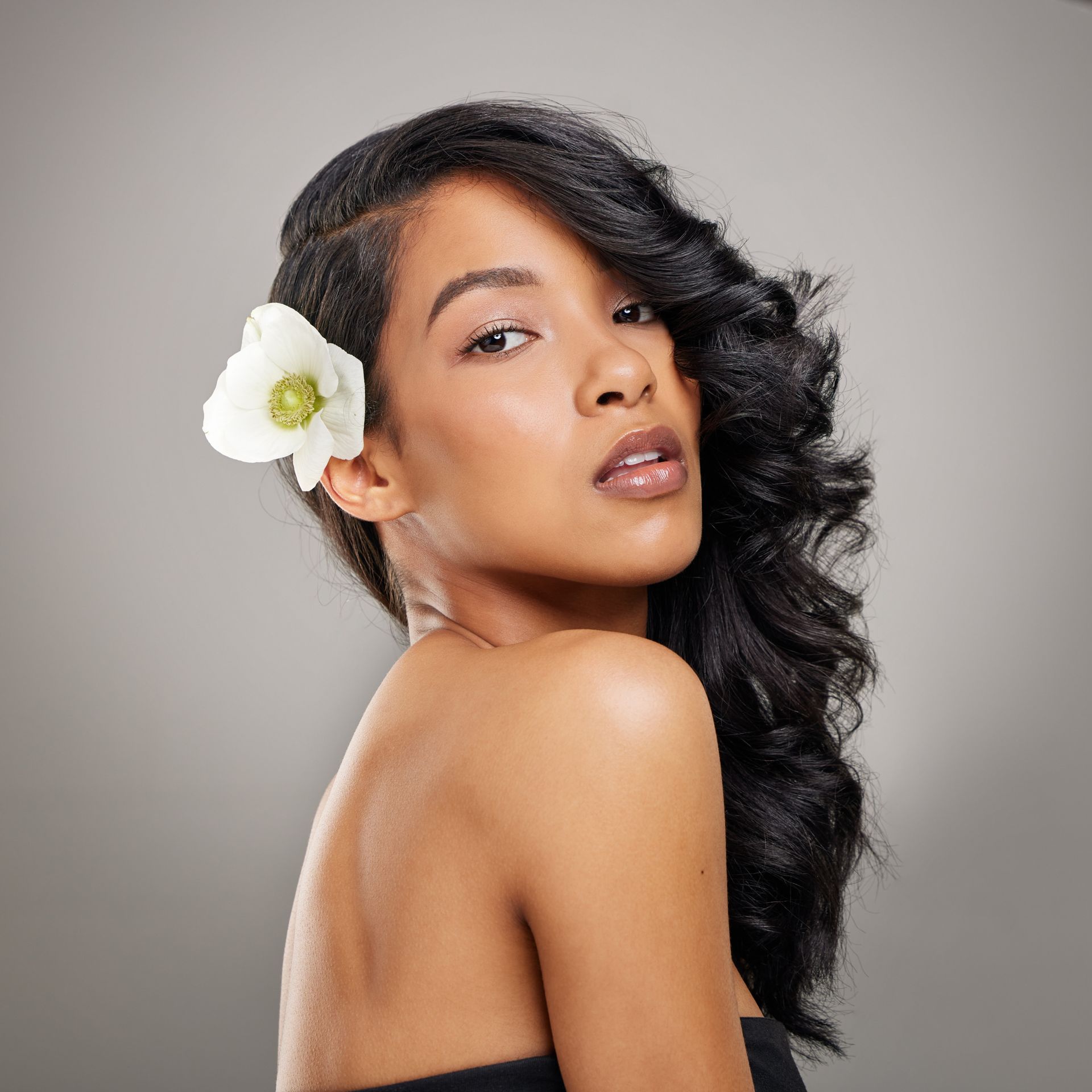Woman with dark wavy hair, wearing a flower, looking over shoulder, neutral background.