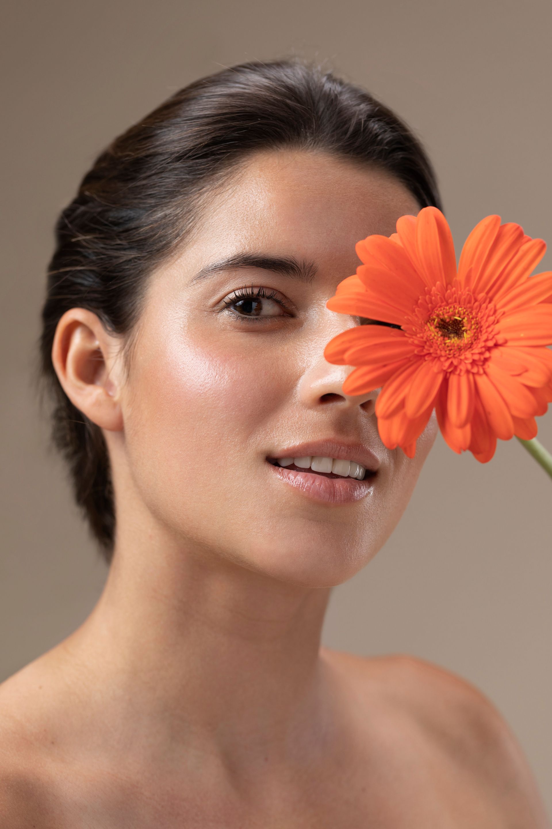 Woman holding a bright orange gerbera daisy near her face, smiling; tan background.