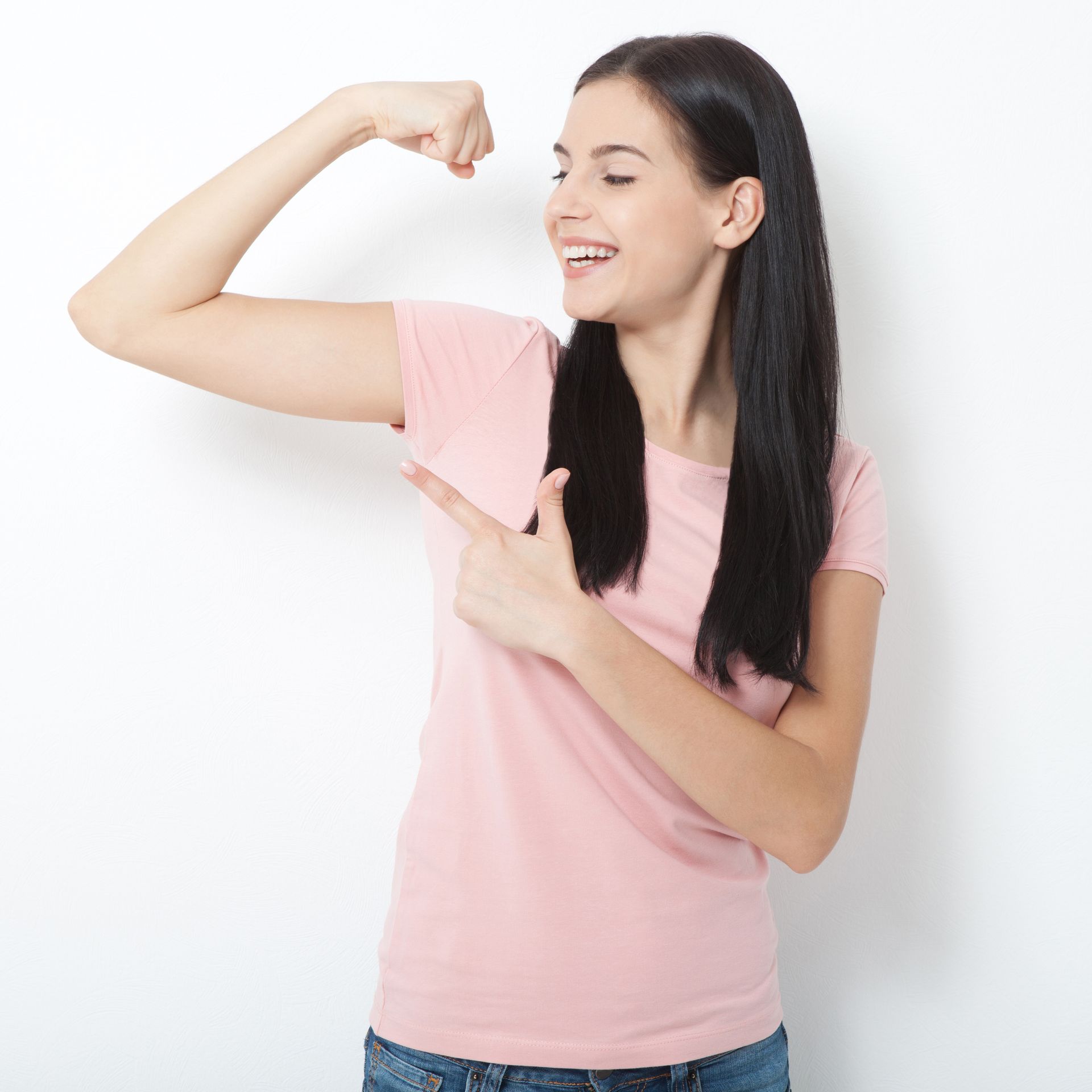 Woman with long dark hair flexing bicep and smiling, pointing at arm. Pink shirt, white background.