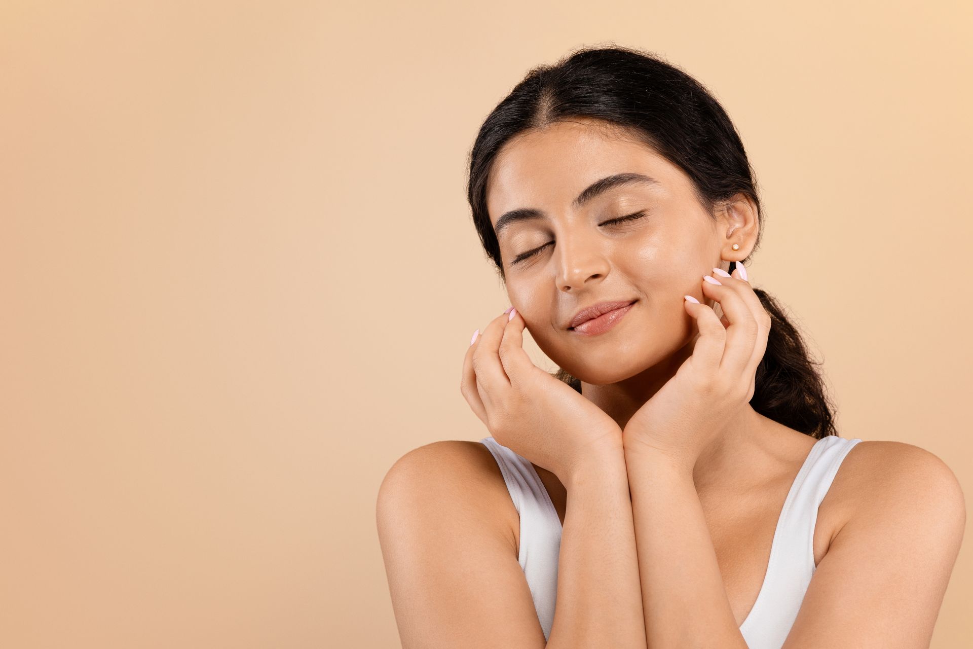 A woman is touching her face with her hands and smiling with her eyes closed.