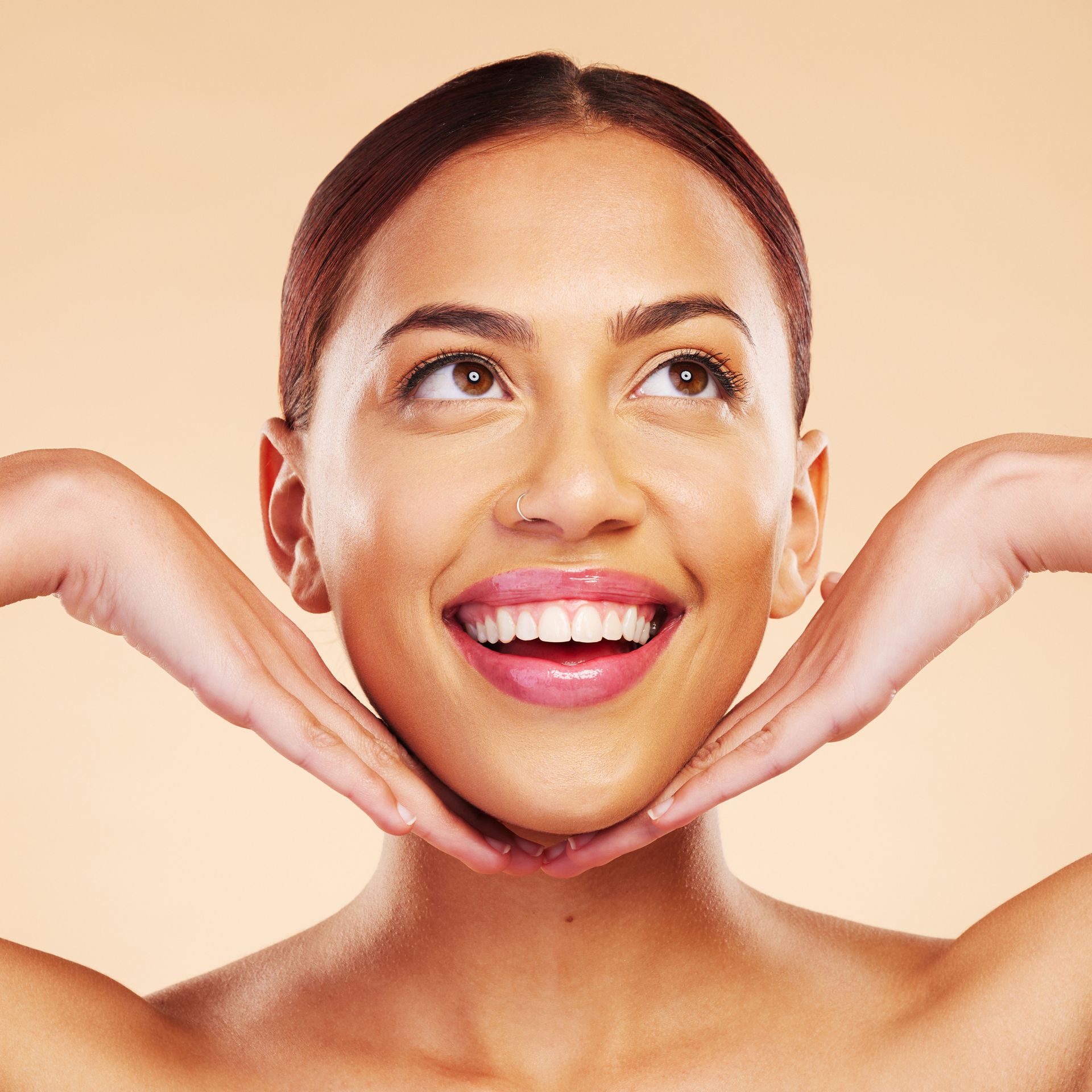 Woman with a bright smile, hands framing her face, looking upwards. Light skin tone, beige background.