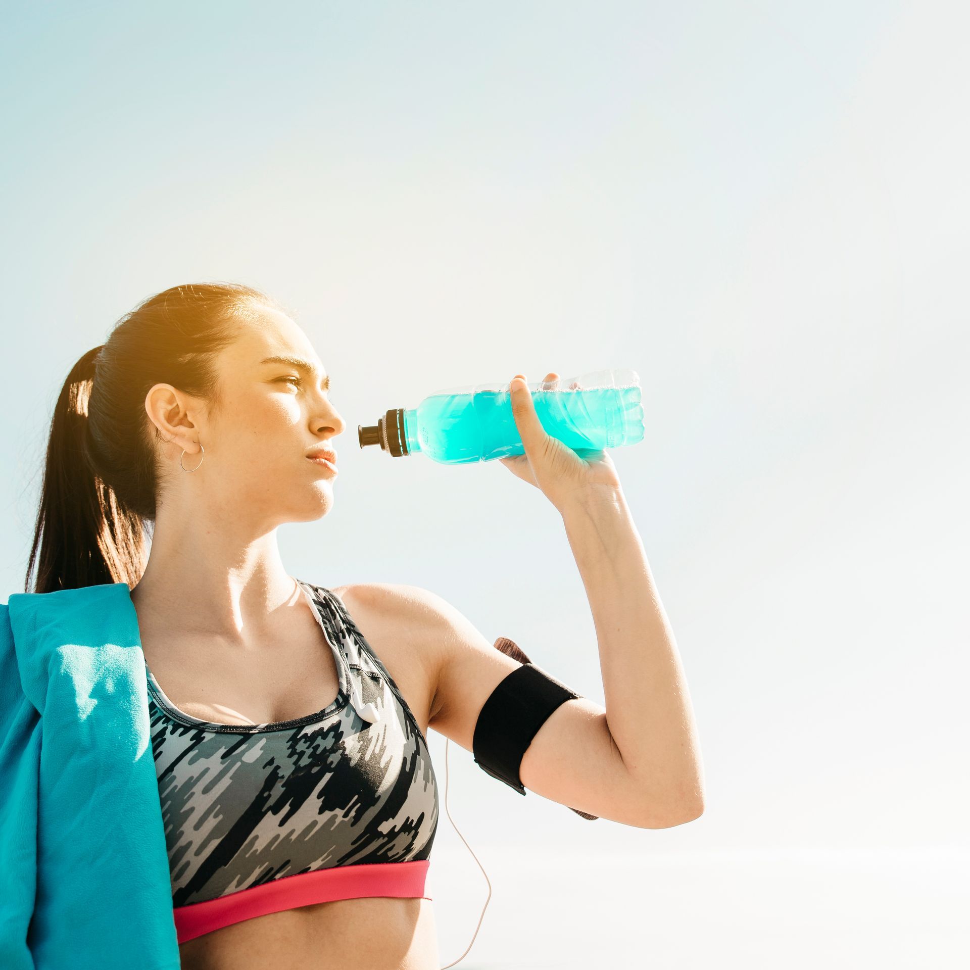 Woman in workout clothes drinking from a blue water bottle with a towel over her shoulder under a bright sky.