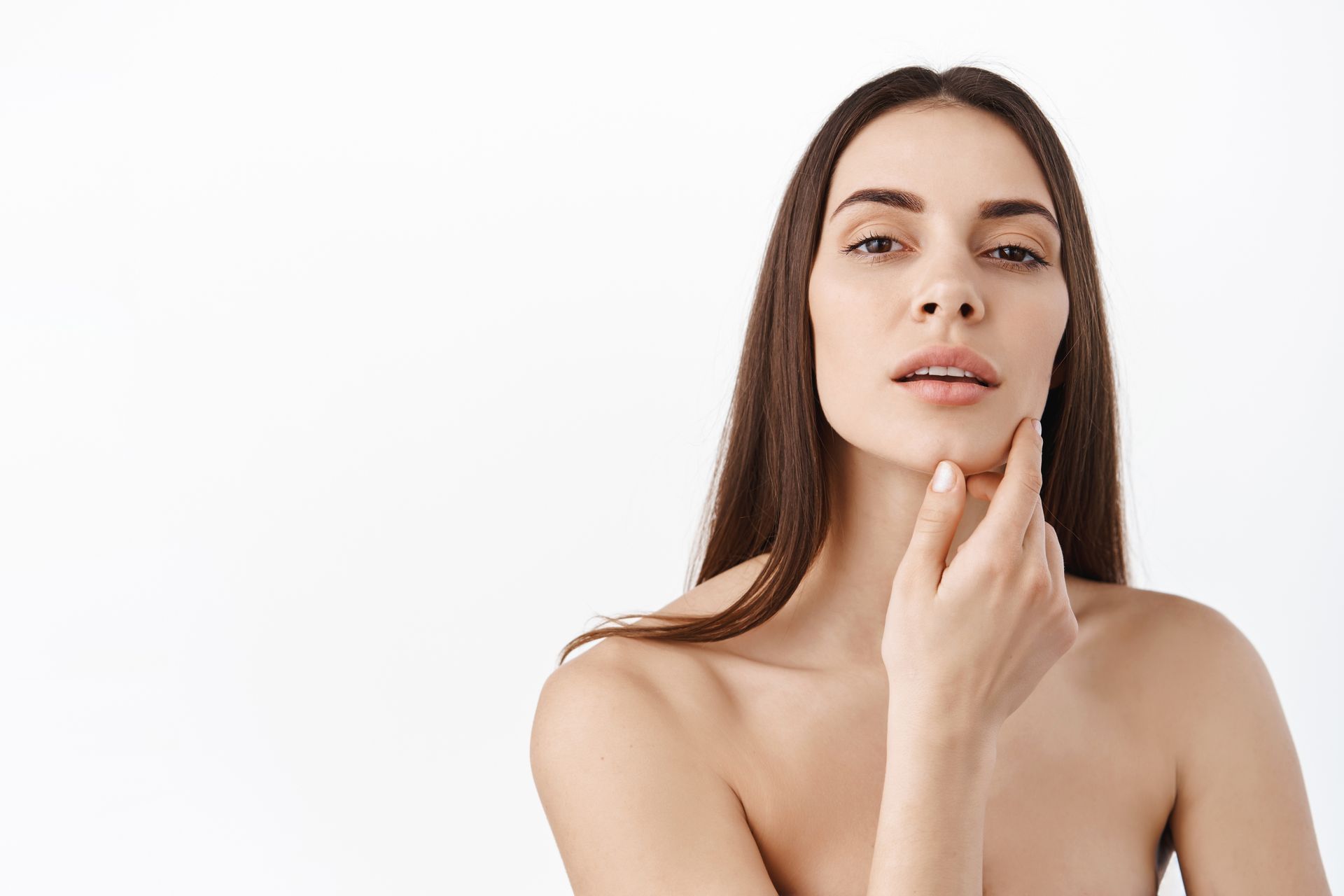 Woman with long brown hair, touching her jaw, looking at the camera, white background.