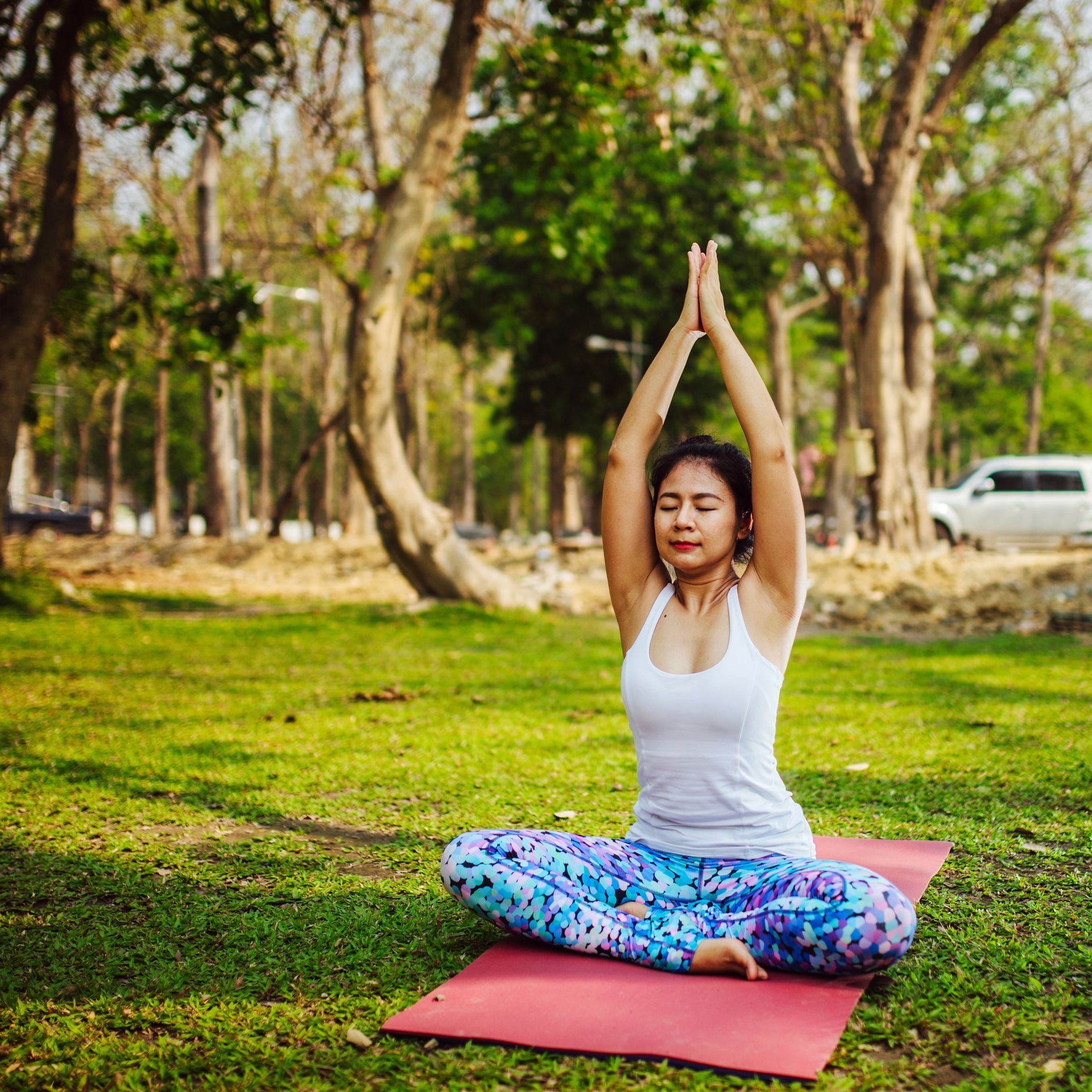 Woman in yoga pose, hands above head, eyes closed, in a park on a pink mat.