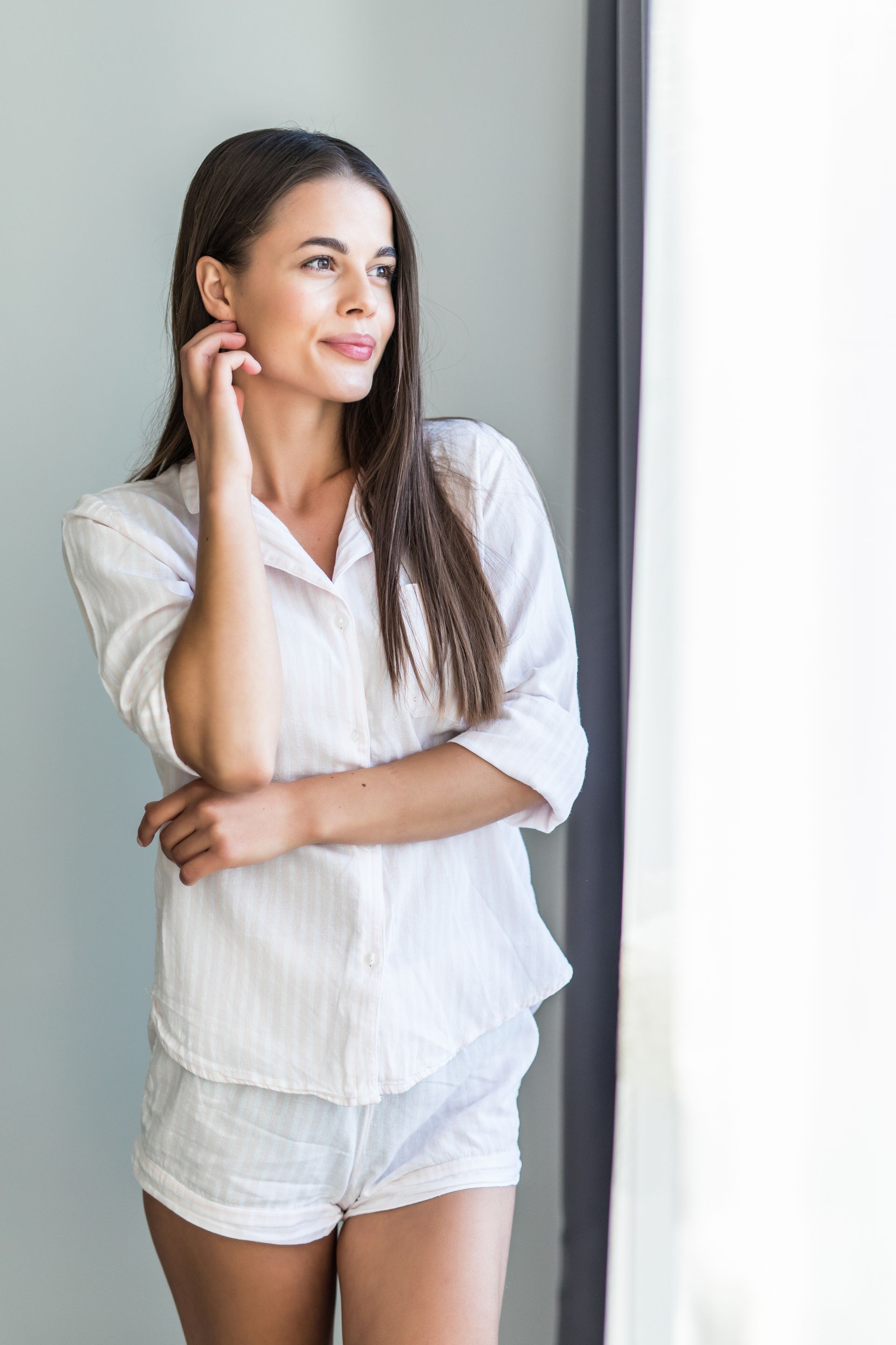 A woman in a white shirt and shorts is standing in front of a window.