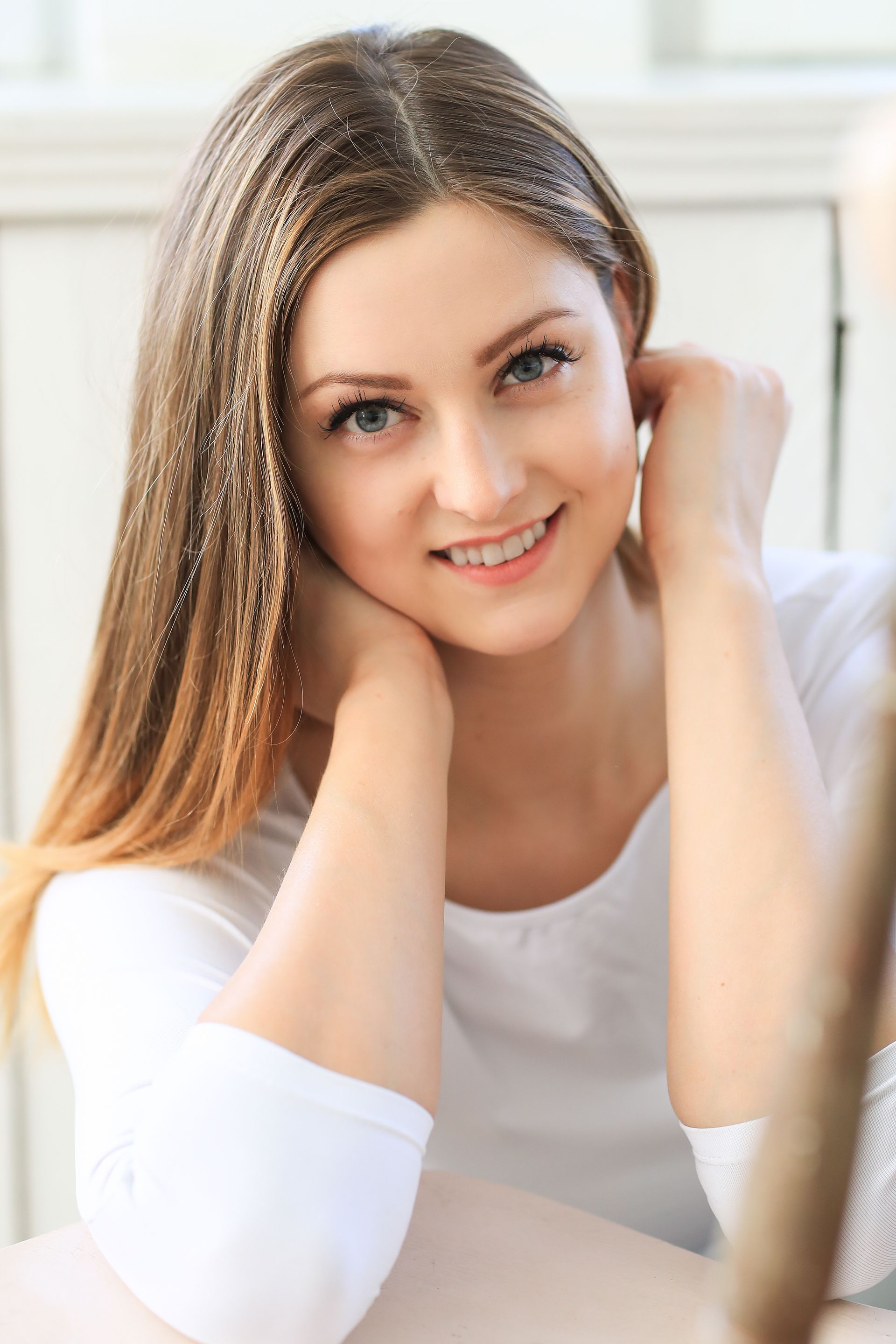 A young woman is sitting at a table with her hands on her face and smiling.