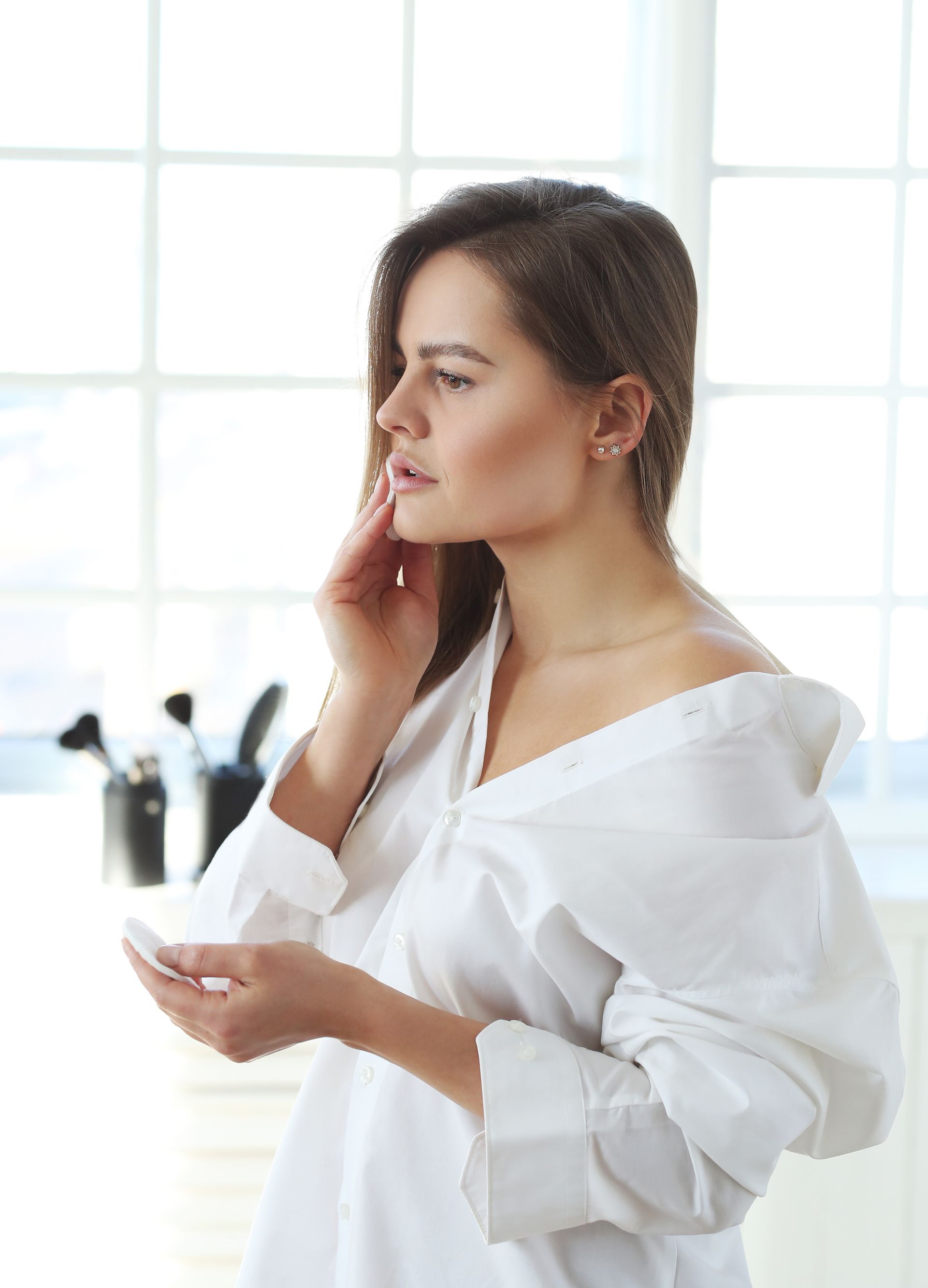 A woman in a white shirt is applying makeup in front of a mirror.