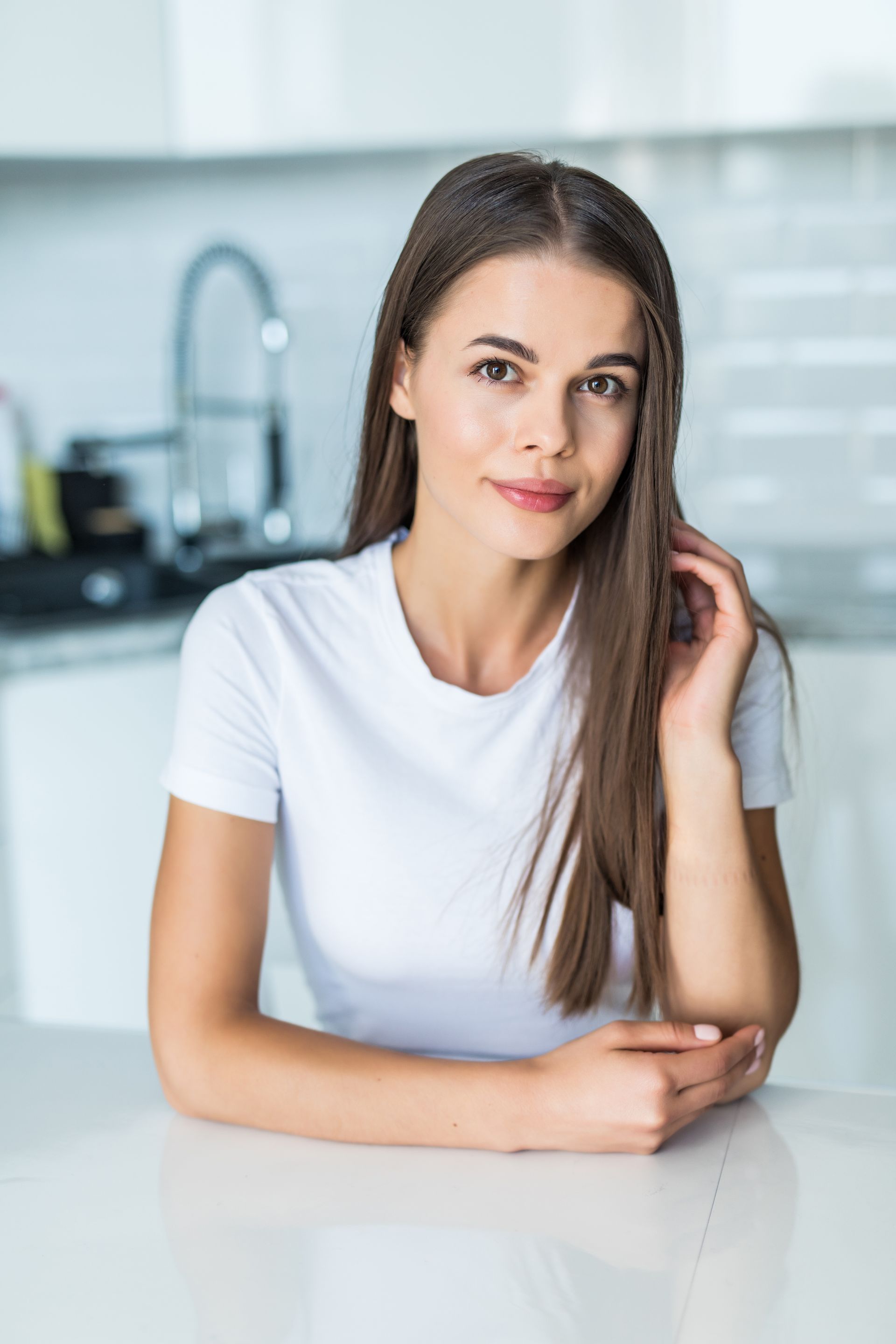 A woman in a white t-shirt is sitting at a table in a kitchen.