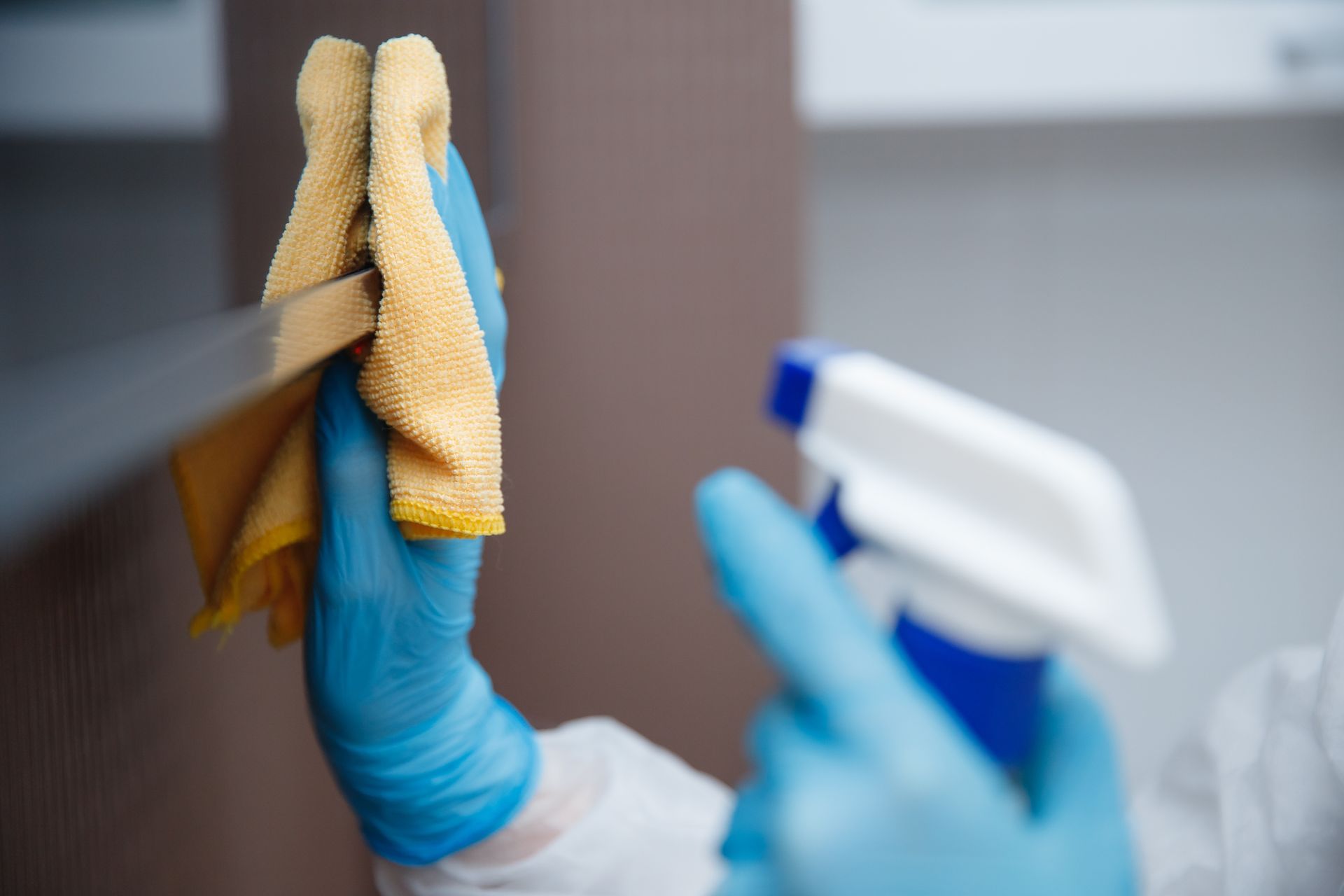 Person in blue gloves spraying cleaner onto a surface and wiping with a yellow cloth.